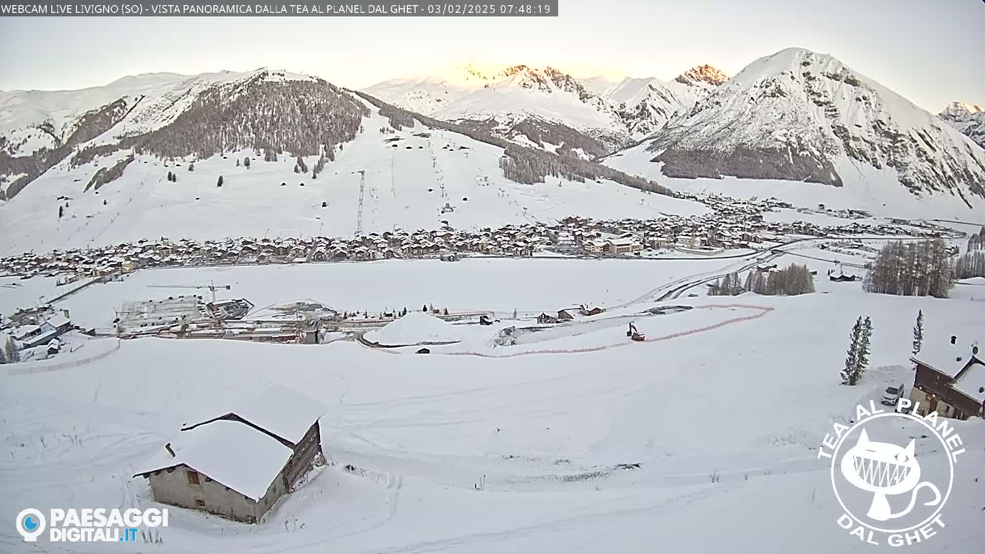 A snow-covered mountain village is nestled in a valley below snow-capped mountains, under a clear, sunrise sky.