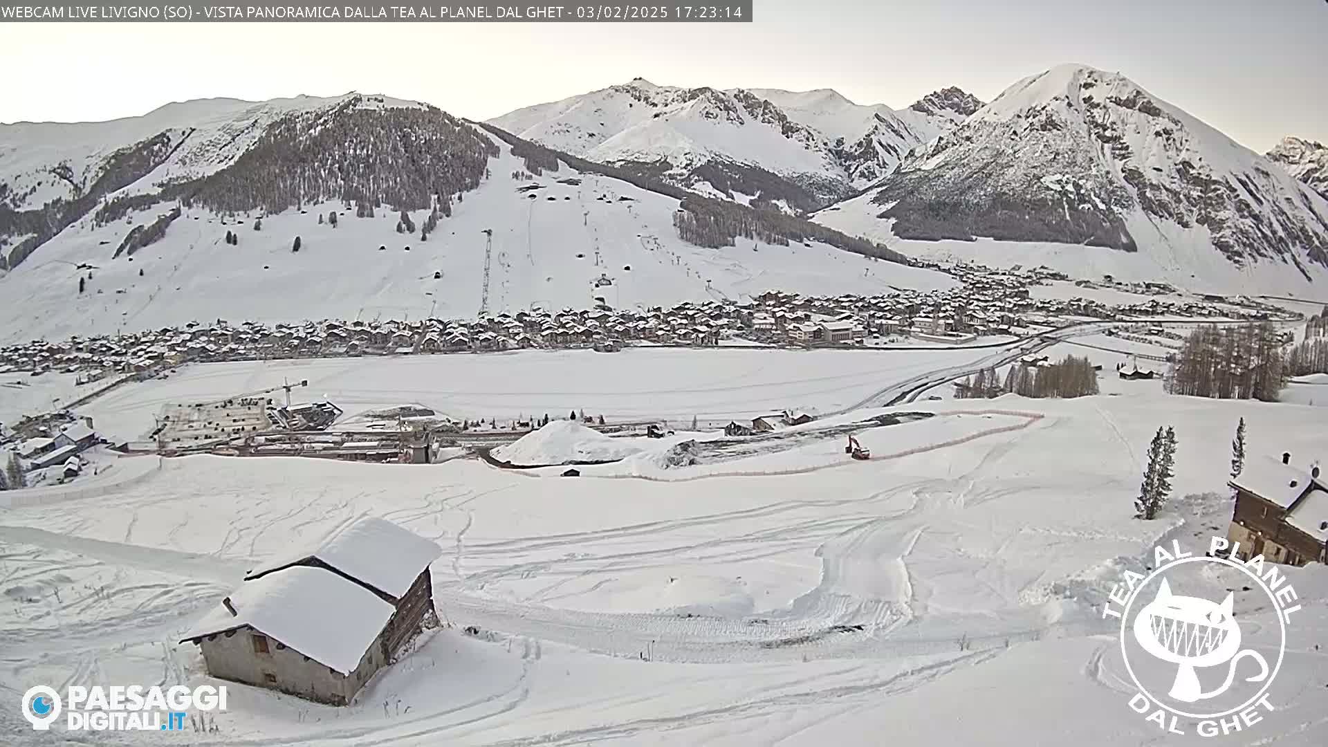 A snow-covered village nestled in a valley between snow-capped mountains, with a small building in the foreground and construction activity visible in the mid-ground.