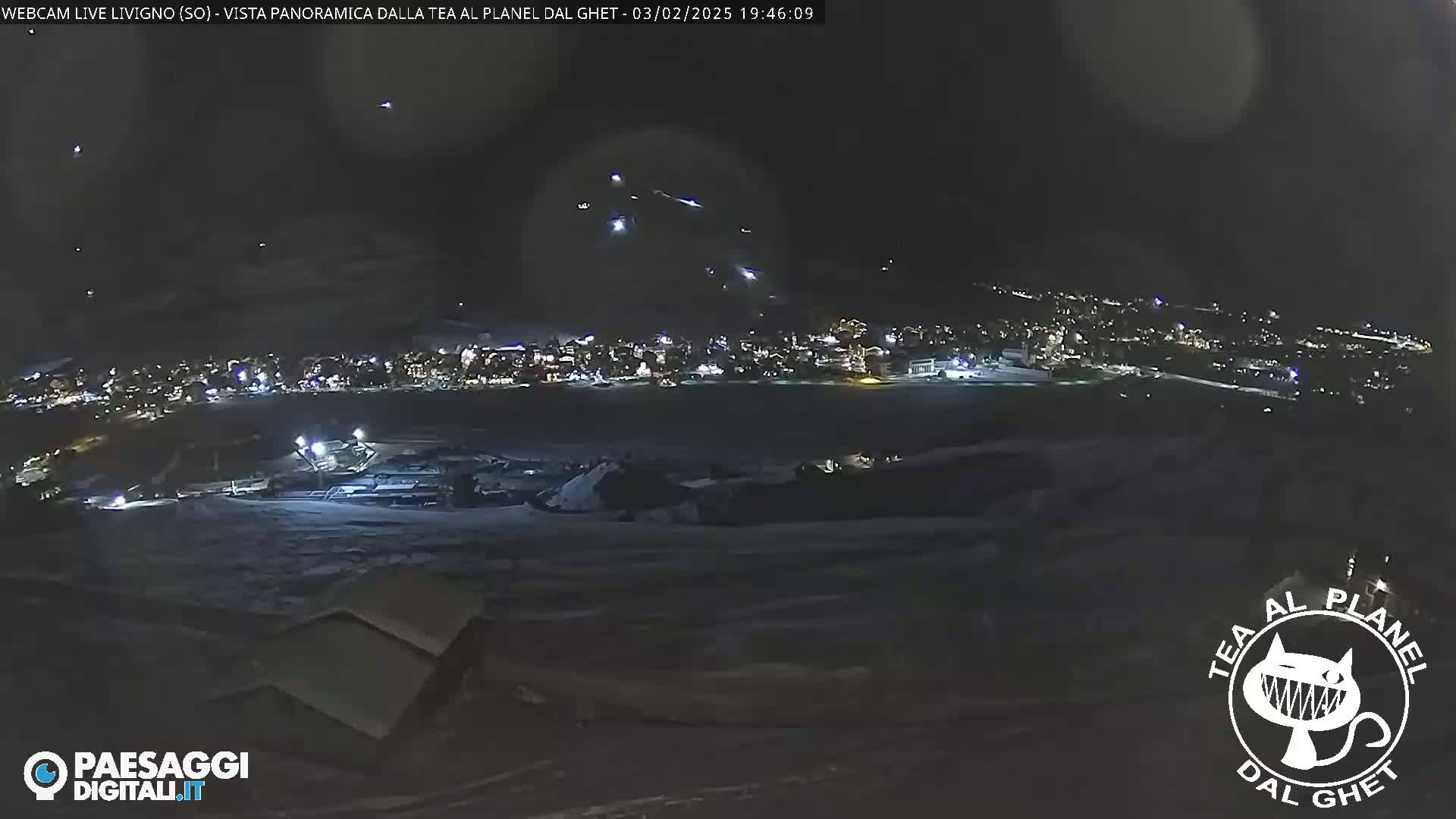 A nighttime long shot shows a snow-covered landscape with a town illuminated in the distance, under a dark sky with several bright lights possibly from aircraft.