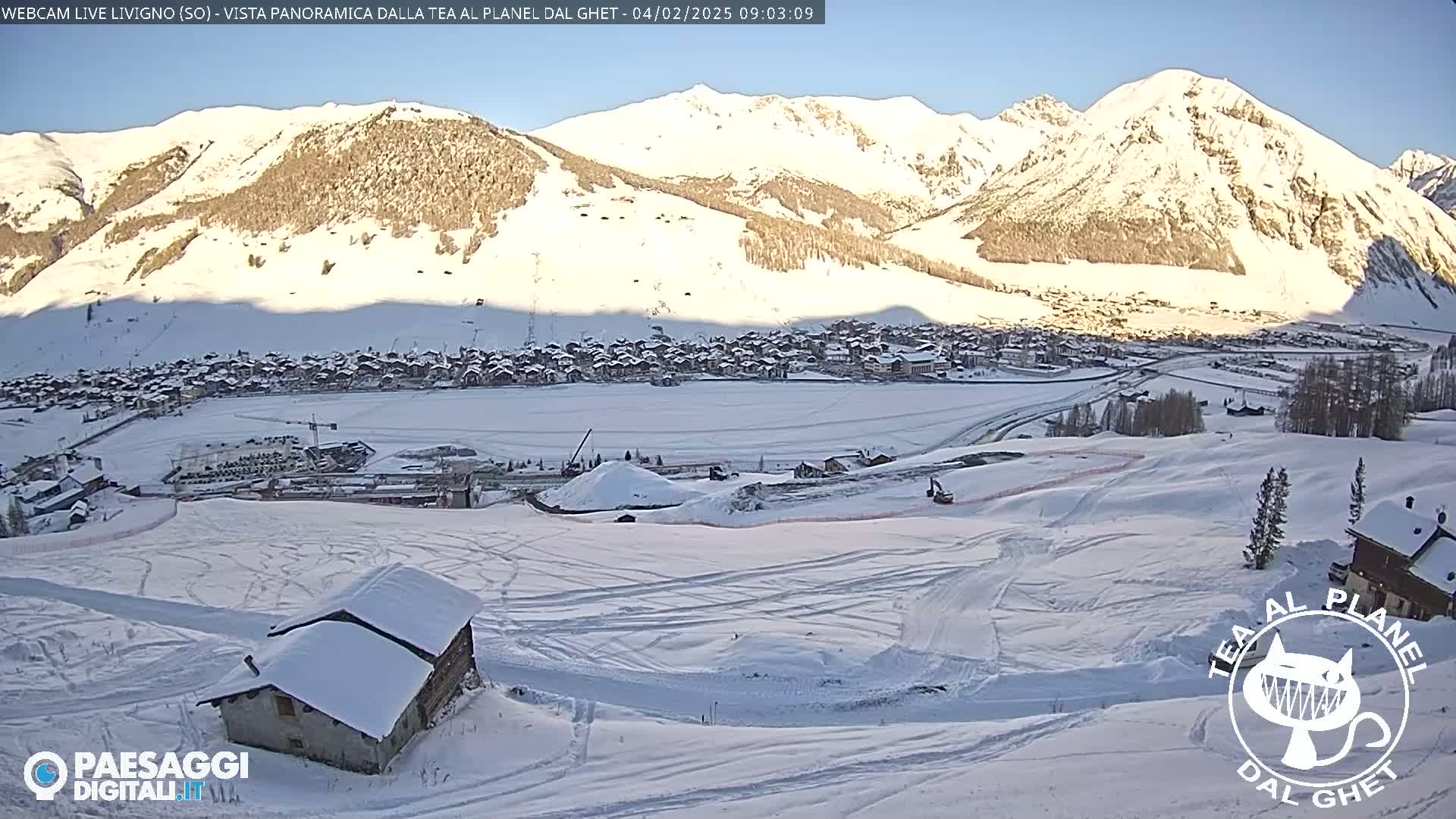 A snow-covered mountain village is nestled below a range of snow-capped mountains under a clear, sunny sky.