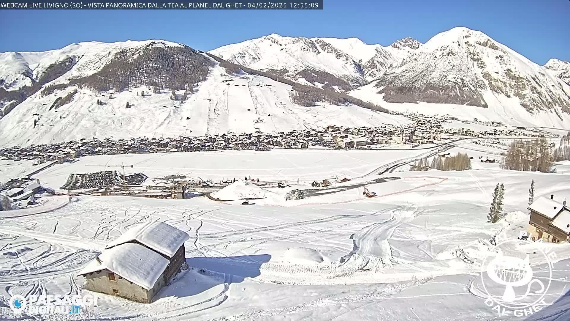 A snow-covered mountain village nestled in a valley under a bright sunny sky, with ski runs and a small building visible in the foreground.