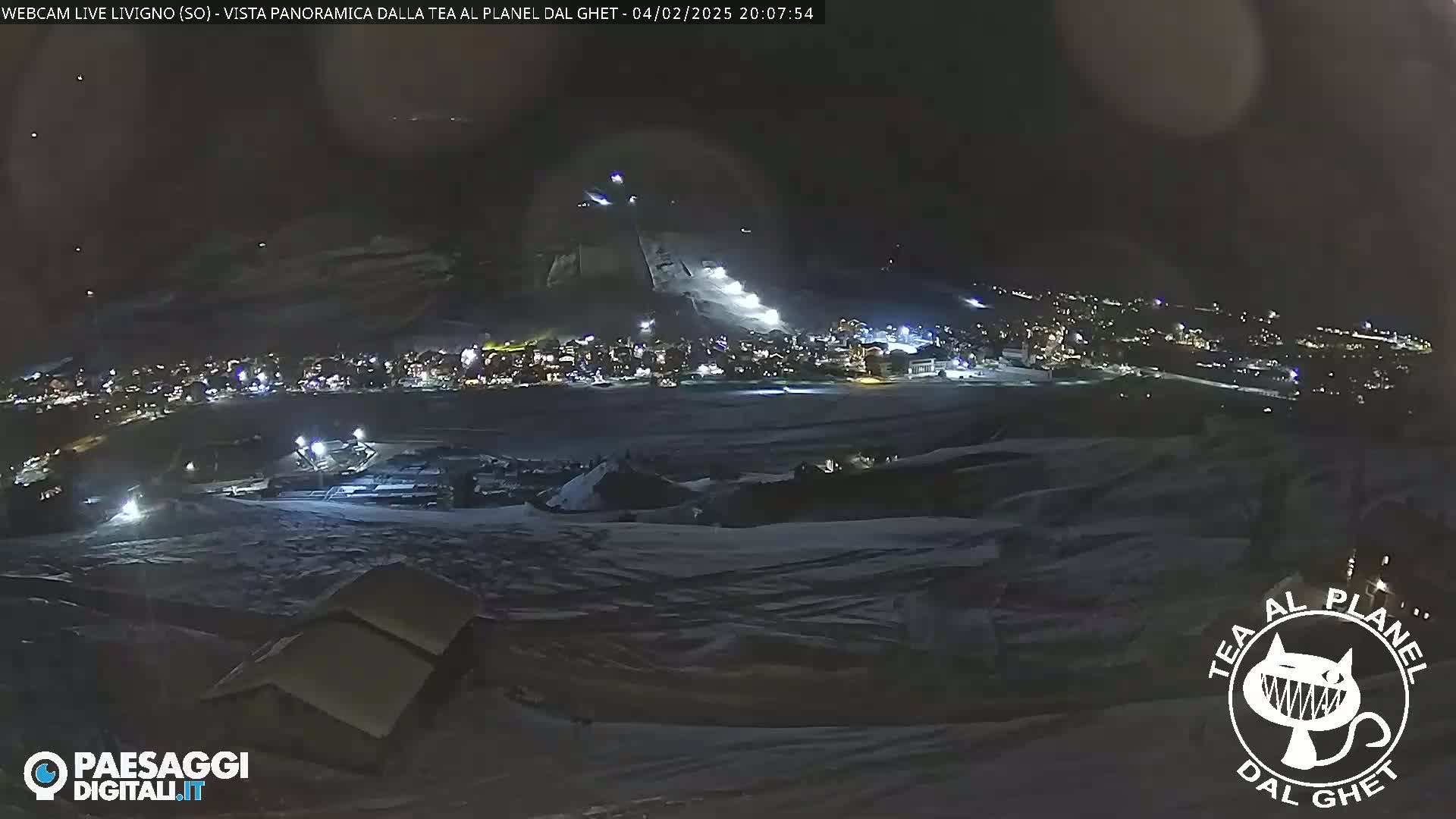 A nighttime, snow-covered landscape shows a brightly lit town below a ski slope.