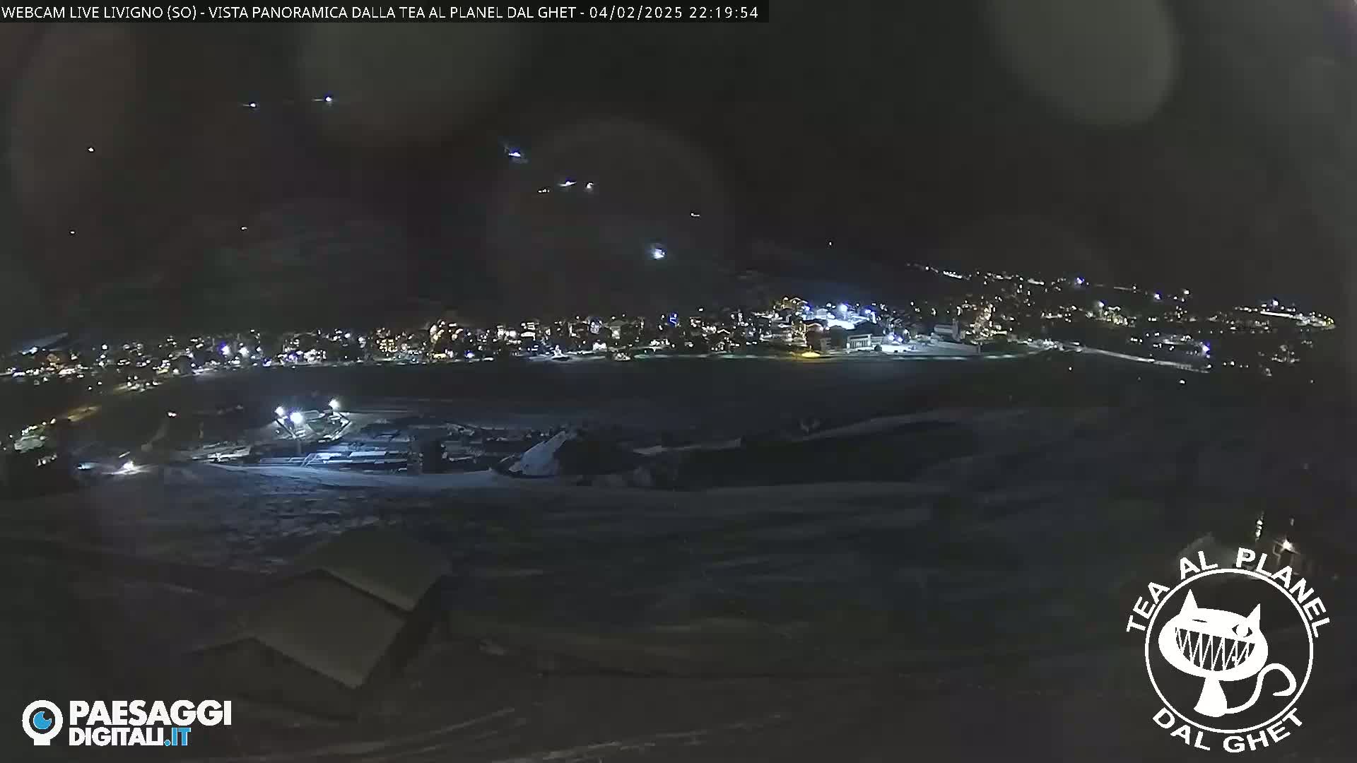 A nighttime, long shot of a snow-covered landscape with a town illuminated in the distance, under a dark sky with several faint lights possibly from aircraft.