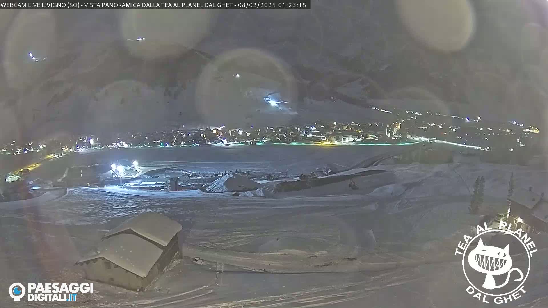 A nighttime, panoramic view of a snow-covered town nestled in a valley, illuminated by lights, under a cloudy sky.