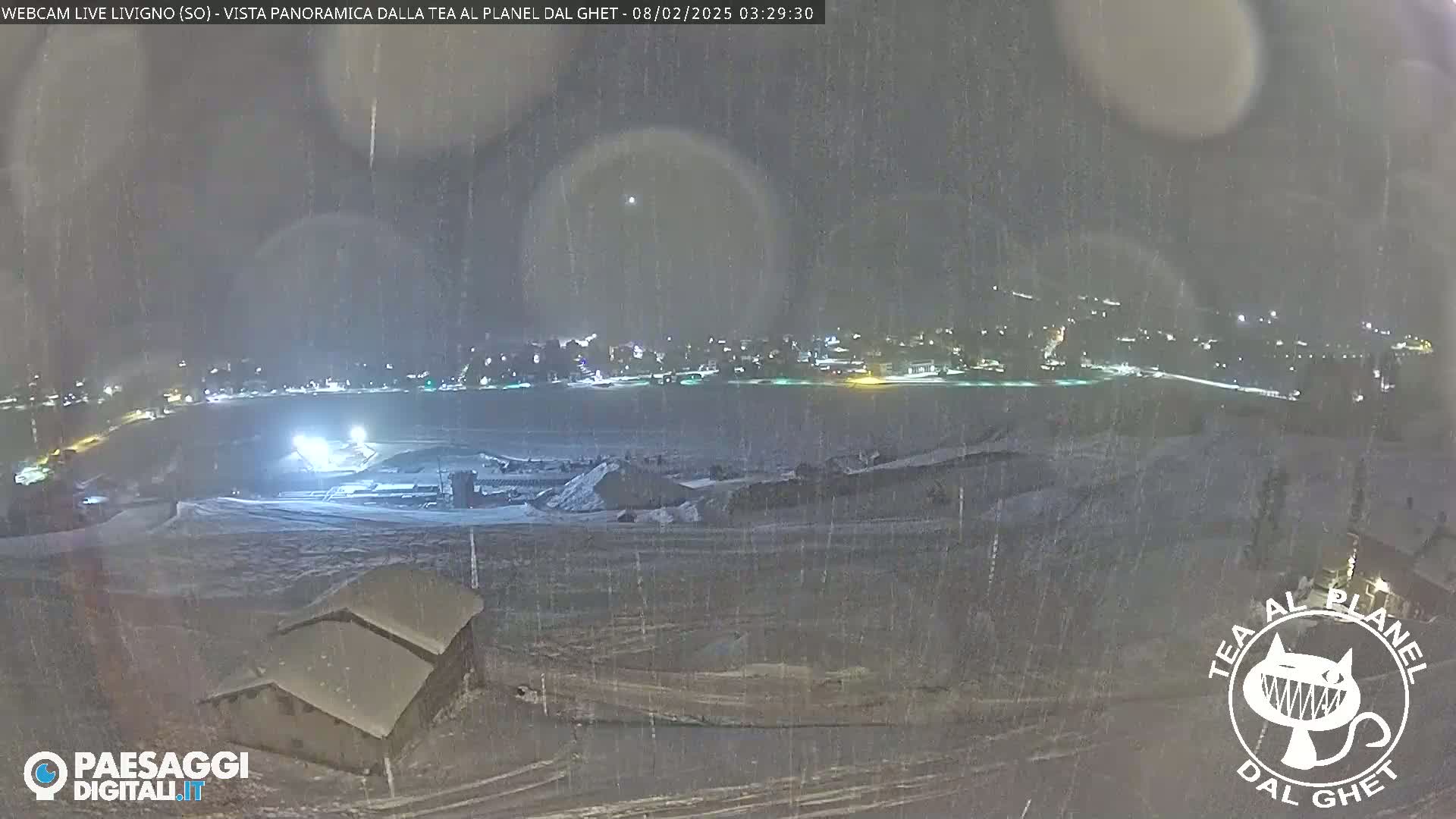 A nighttime, snowy scene shows a snow-covered landscape with a small building in the foreground and a town illuminated in the distance under falling snow.