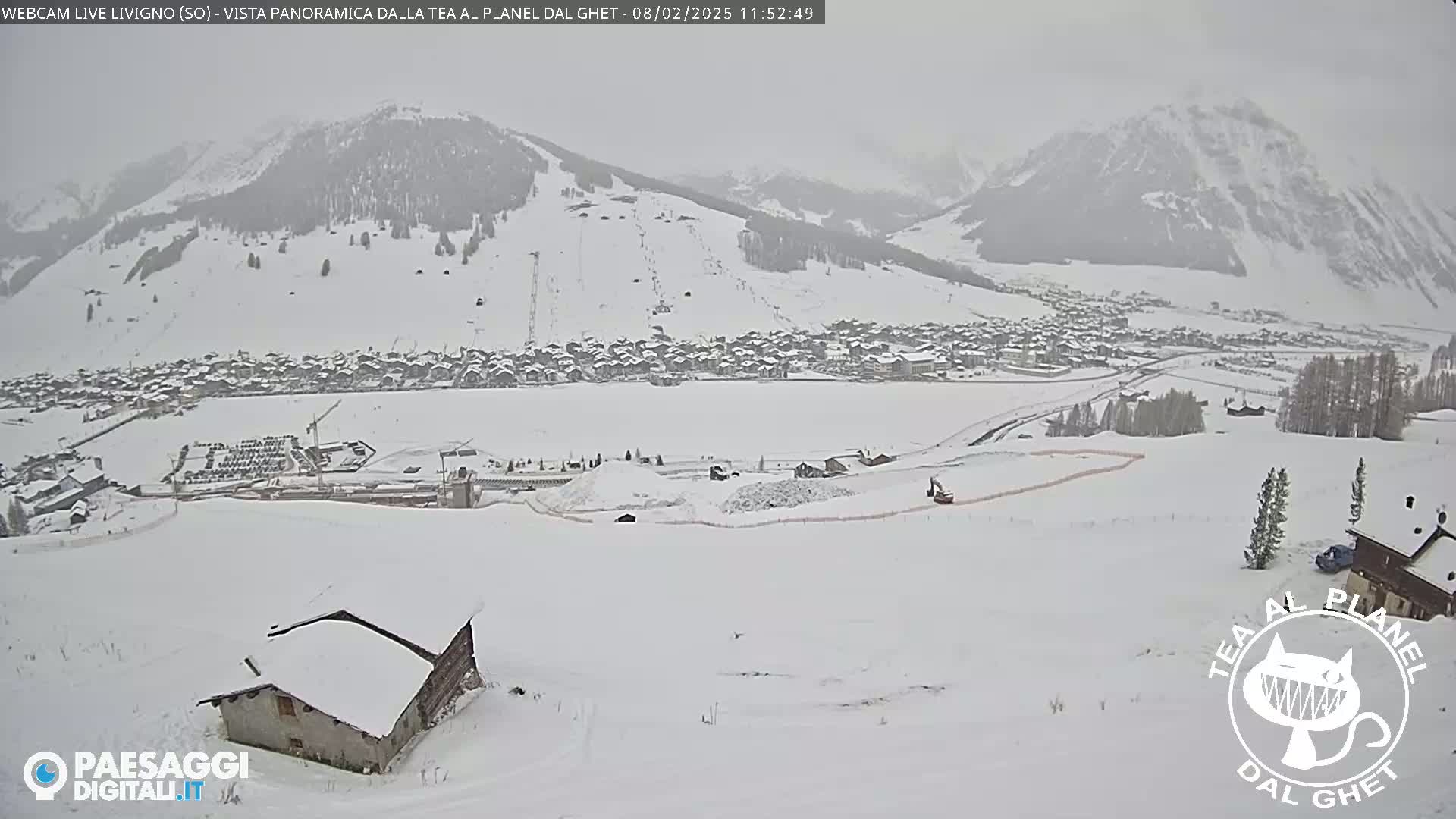 A snow-covered landscape reveals a village nestled in a valley beneath snow-dusted mountains, under an overcast sky.