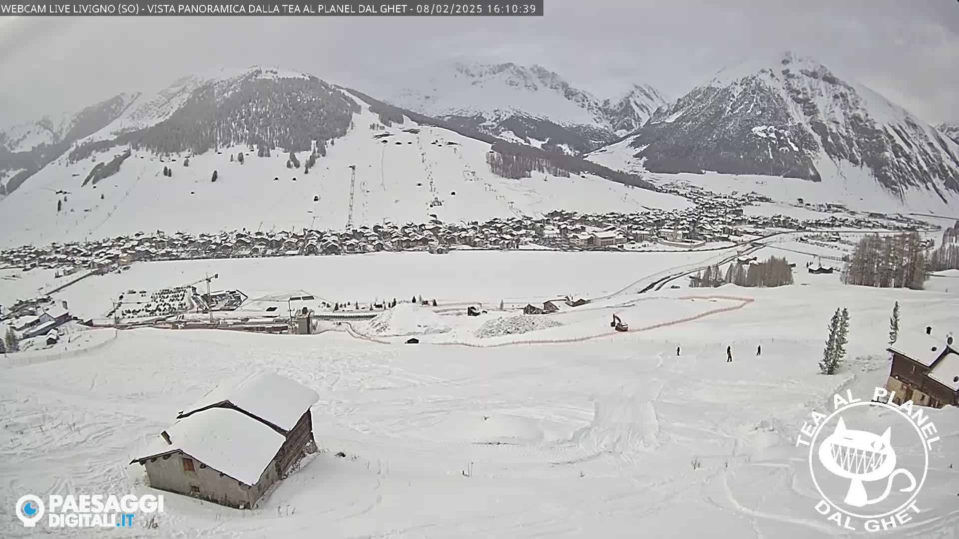 A snow-covered mountain village is viewed from a snowy hillside, with overcast skies and several people visible on a ski slope.