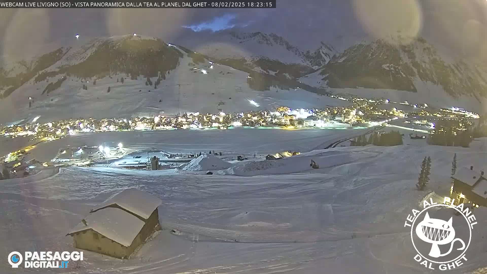 A snow-covered landscape at night, showing a village illuminated below a mountain, with a small building in the foreground.
