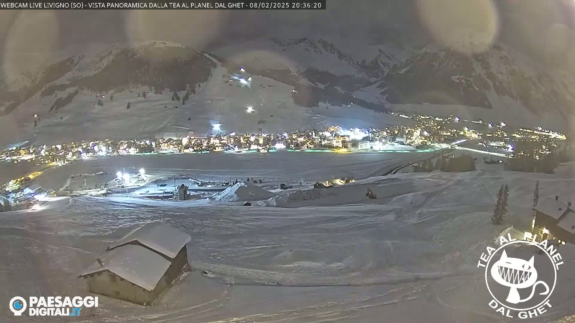 A snow-covered mountain village at night, illuminated by lights, is viewed from a high vantage point overlooking snow-covered slopes.