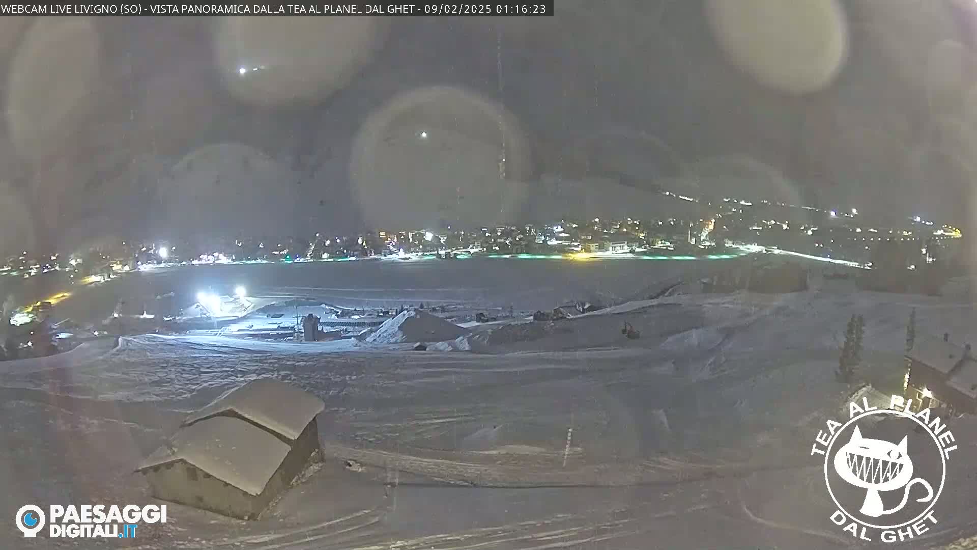 A snow-covered landscape at night, showing a town in the distance illuminated by lights, under a dark, slightly hazy sky.