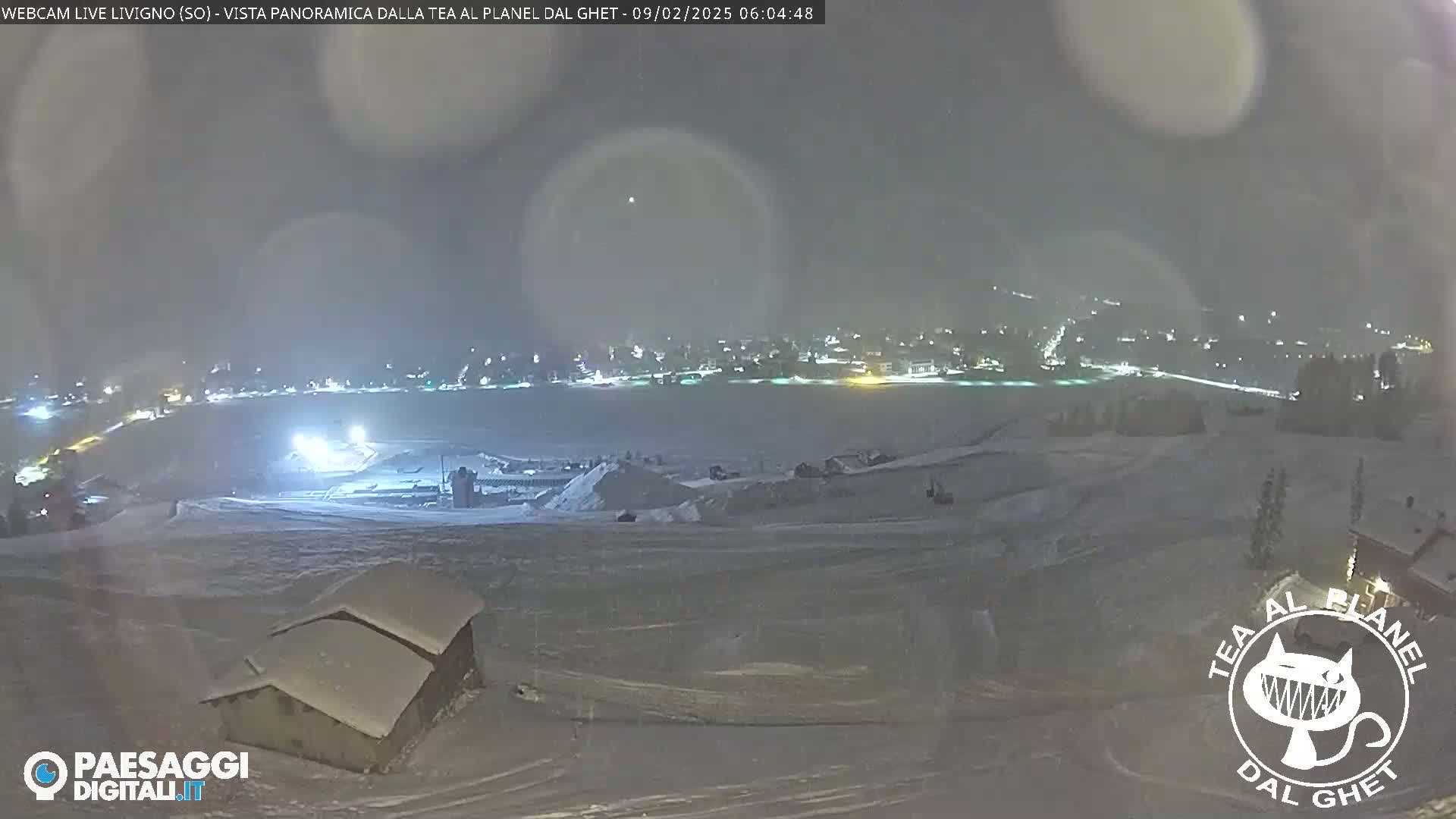 A snow-covered landscape at night shows a town in the distance illuminated by lights, with a small snow-covered building in the foreground under a hazy sky.