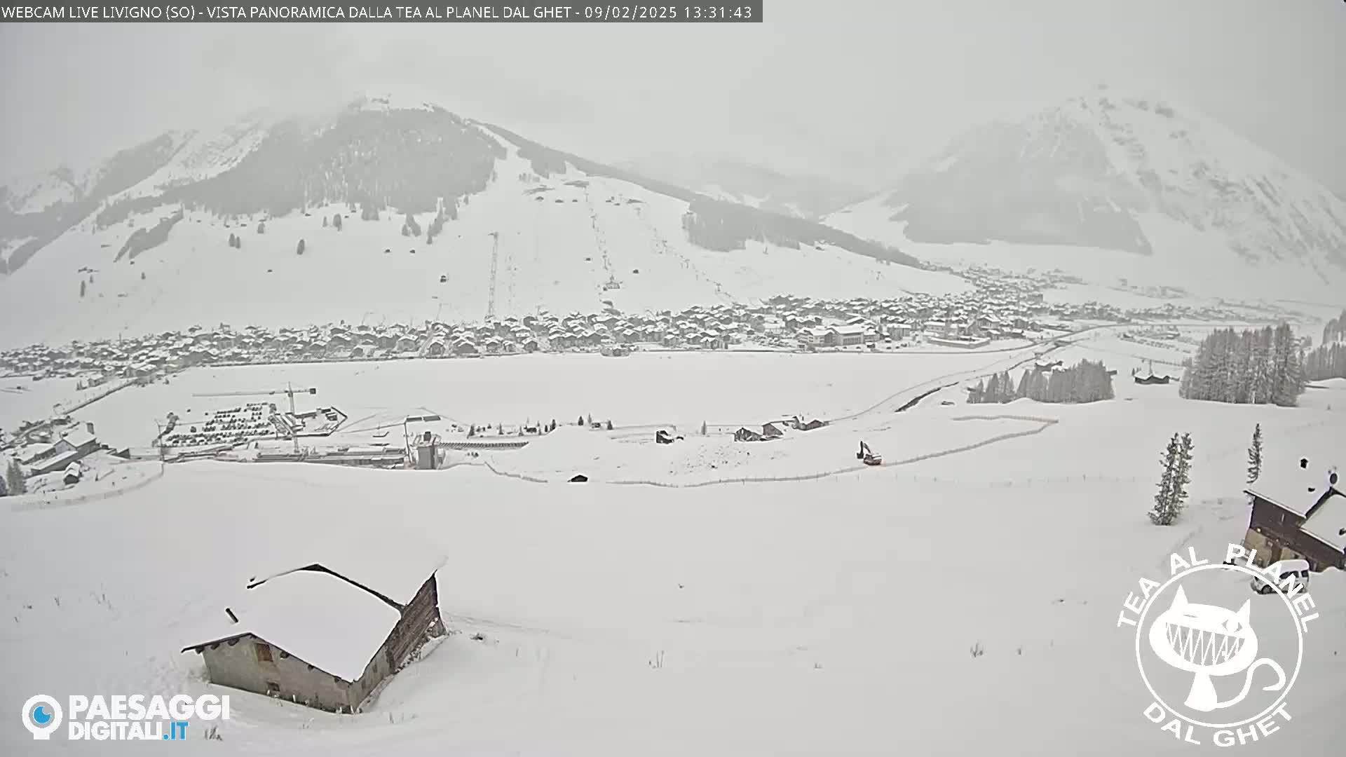 A snow-covered village nestled in a valley below a snow-dusted mountain range, under a hazy, snowy sky.