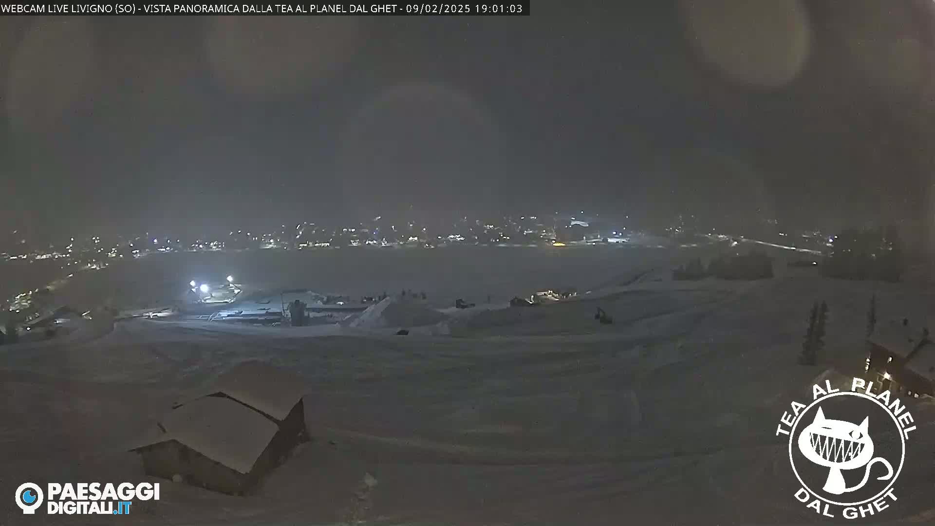 A nighttime, snowy landscape shows a town's lights in the distance, with a snow-covered foreground containing some structures.