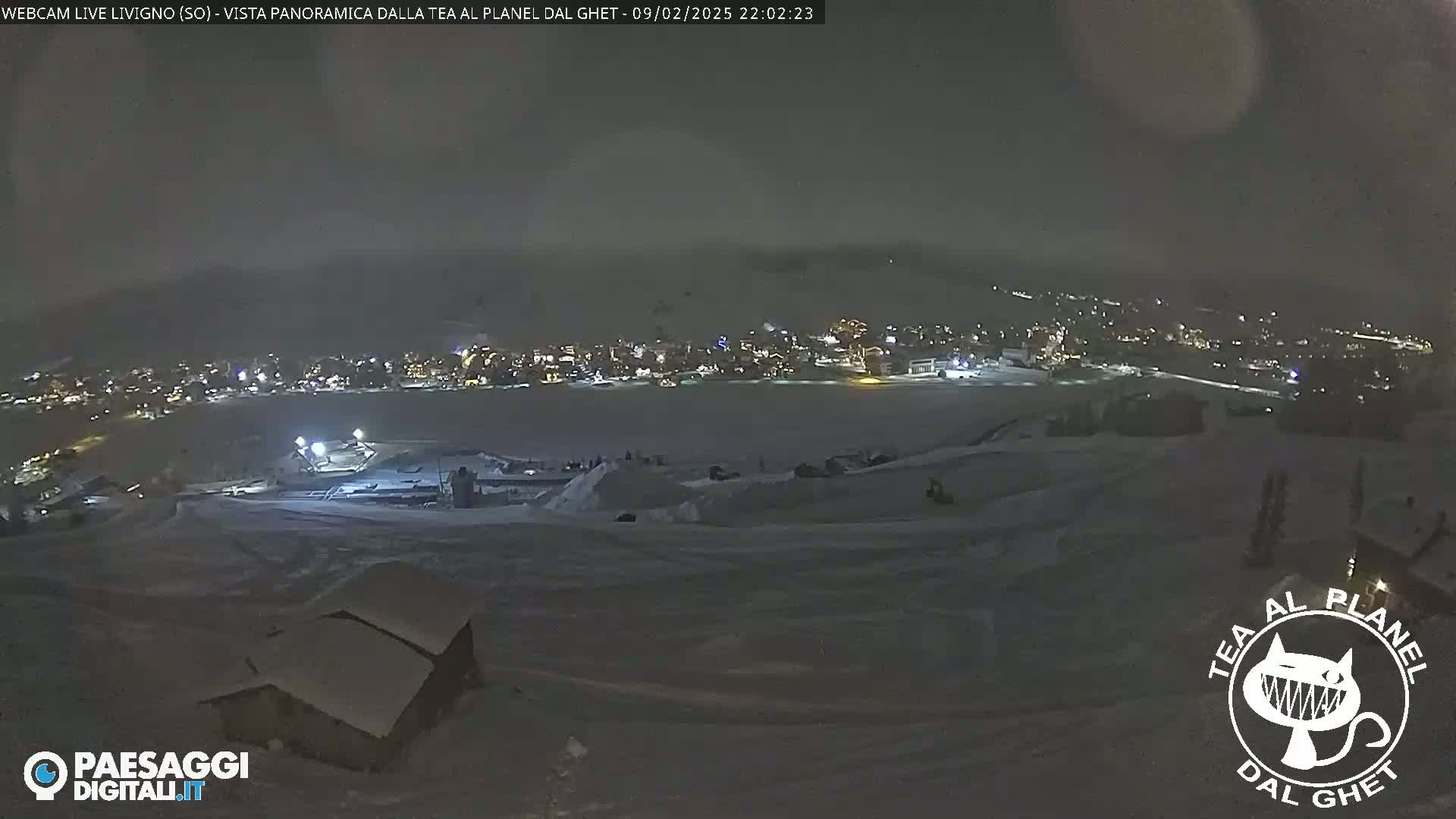 A nighttime, snow-covered landscape shows a town with illuminated buildings in the distance, under a dark, slightly hazy sky.