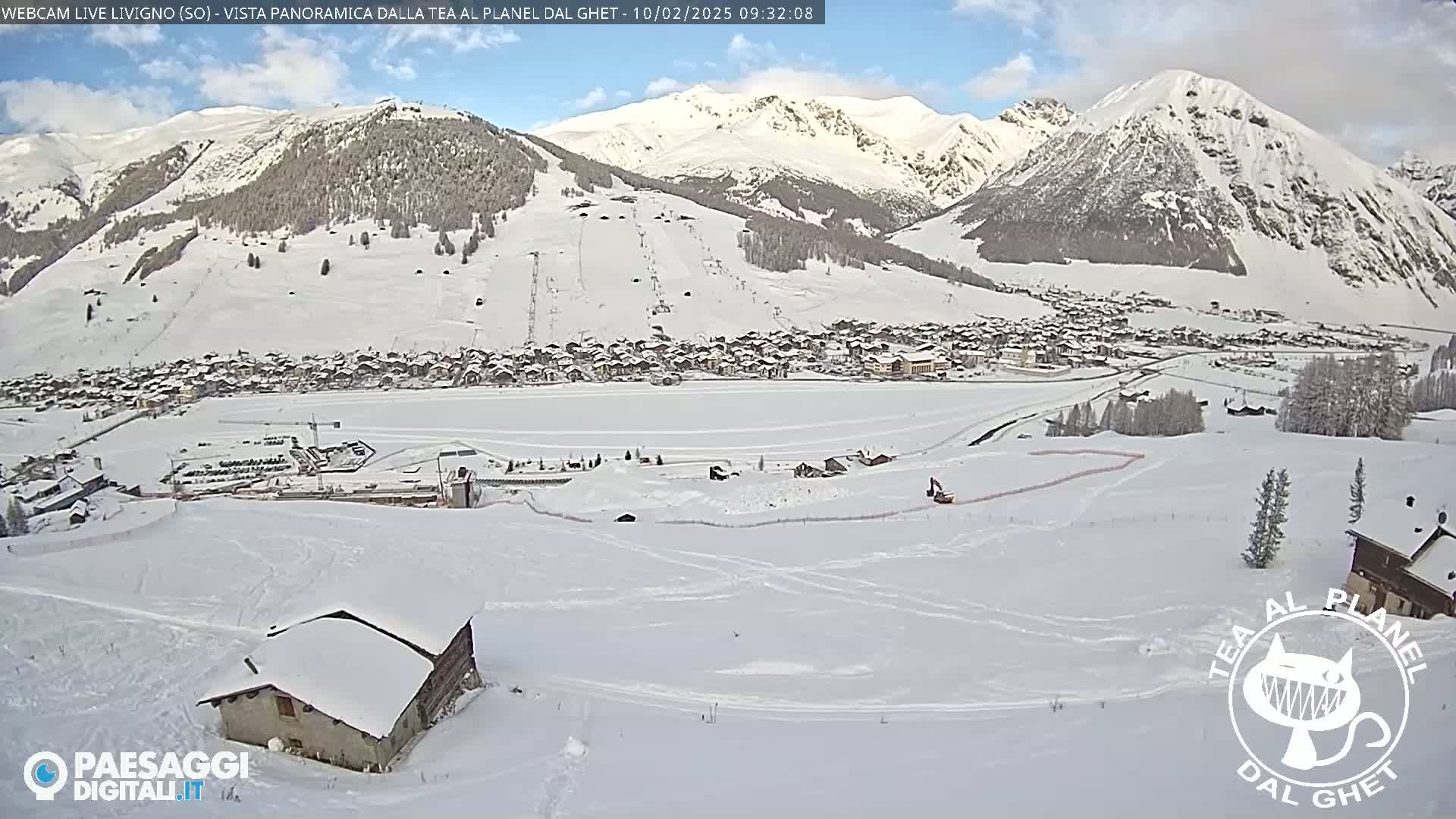 A snow-covered mountain village is nestled in a valley, with snow-capped mountains in the background under a partly cloudy sky.