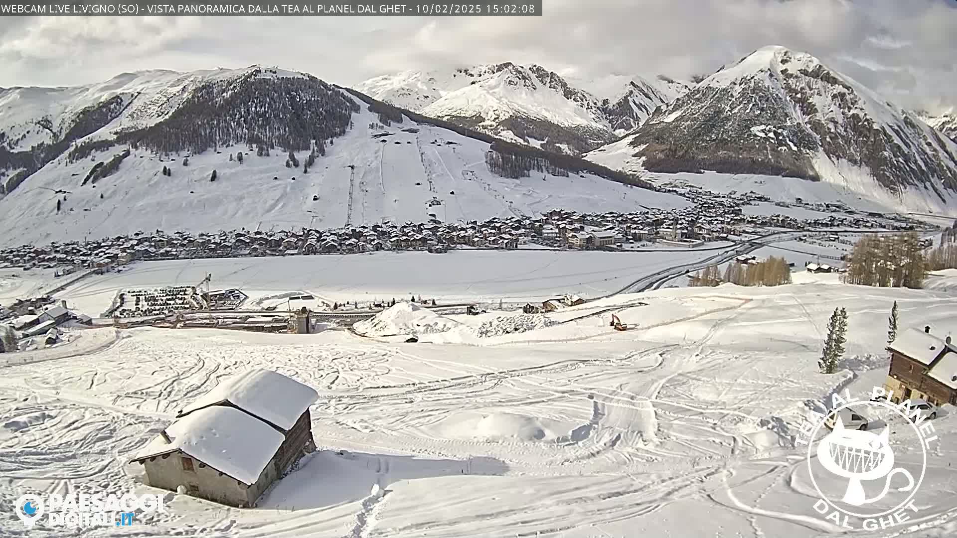 A snow-covered village nestled in a valley between snow-capped mountains under a partly cloudy sky.