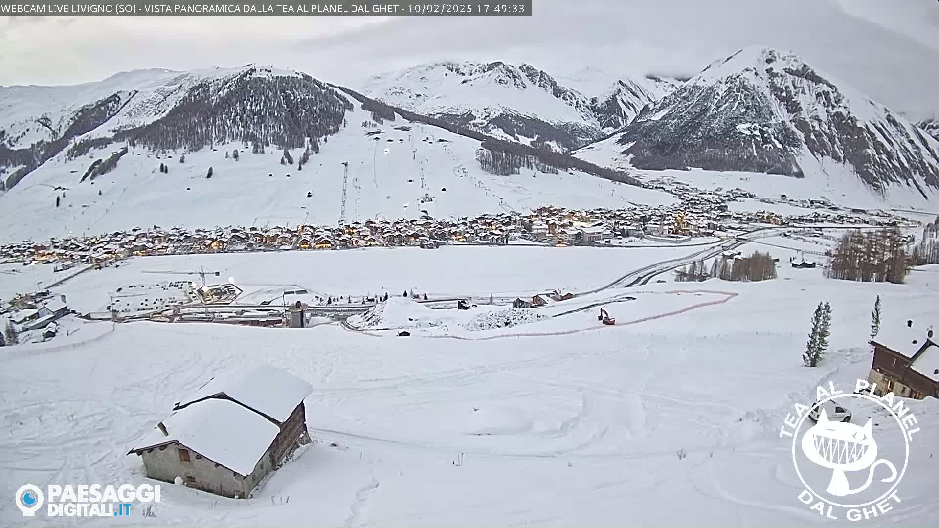 A snow-covered mountain village is nestled in a valley, under an overcast sky; a small snow-covered building is visible in the foreground.