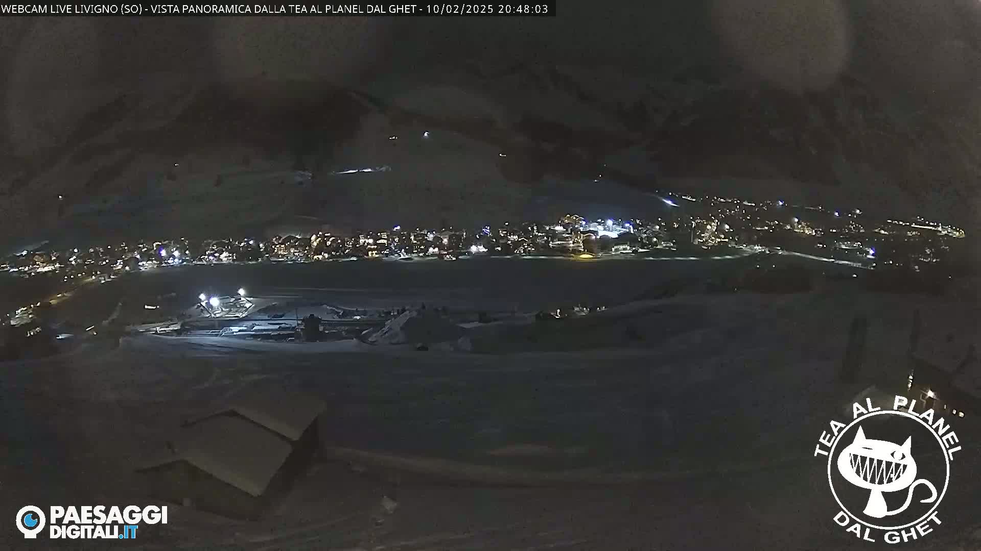 A nighttime, snowy, panoramic view of a town illuminated with lights, nestled in a valley surrounded by mountains.