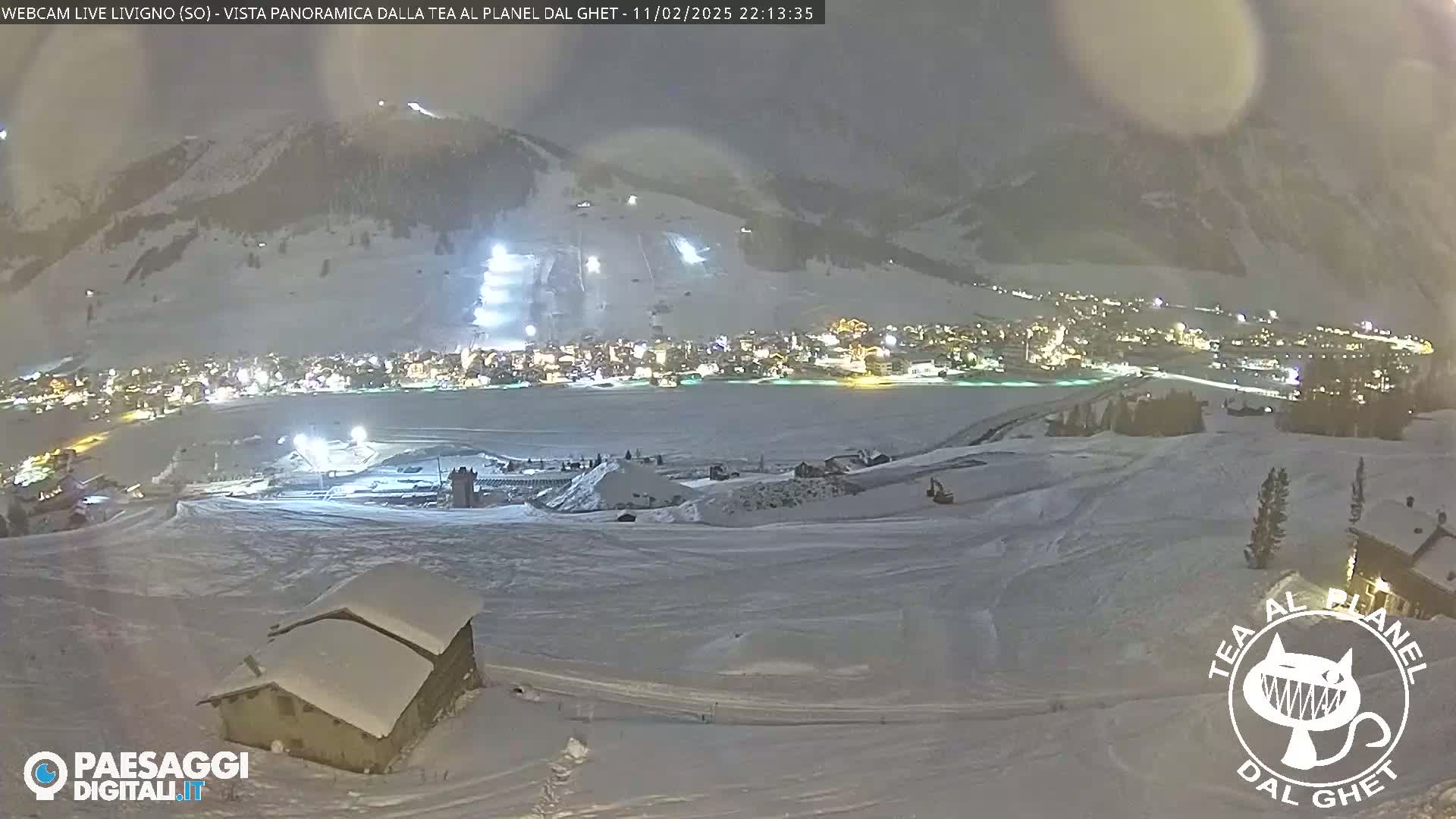 A snow-covered landscape at night, showing a village illuminated below with a mountain in the background under a cloudy sky.