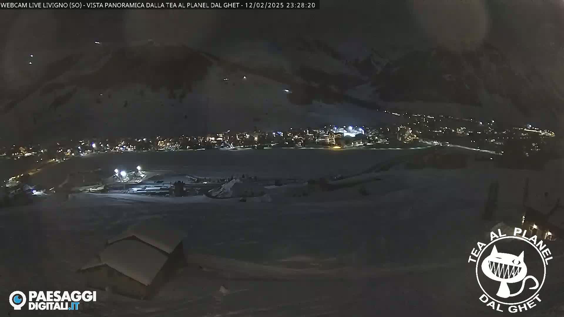 A nighttime, snow-covered landscape shows a town with illuminated buildings in the distance, a small structure in the foreground, and dark, mountainous terrain under a mostly clear sky.