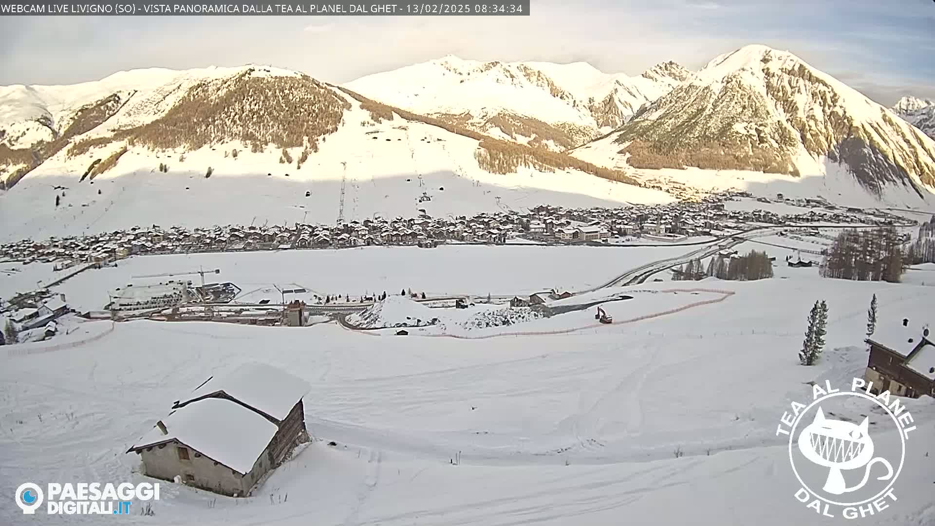 A snow-covered mountain village is nestled in a valley below snow-capped mountains under a mostly clear sky.
