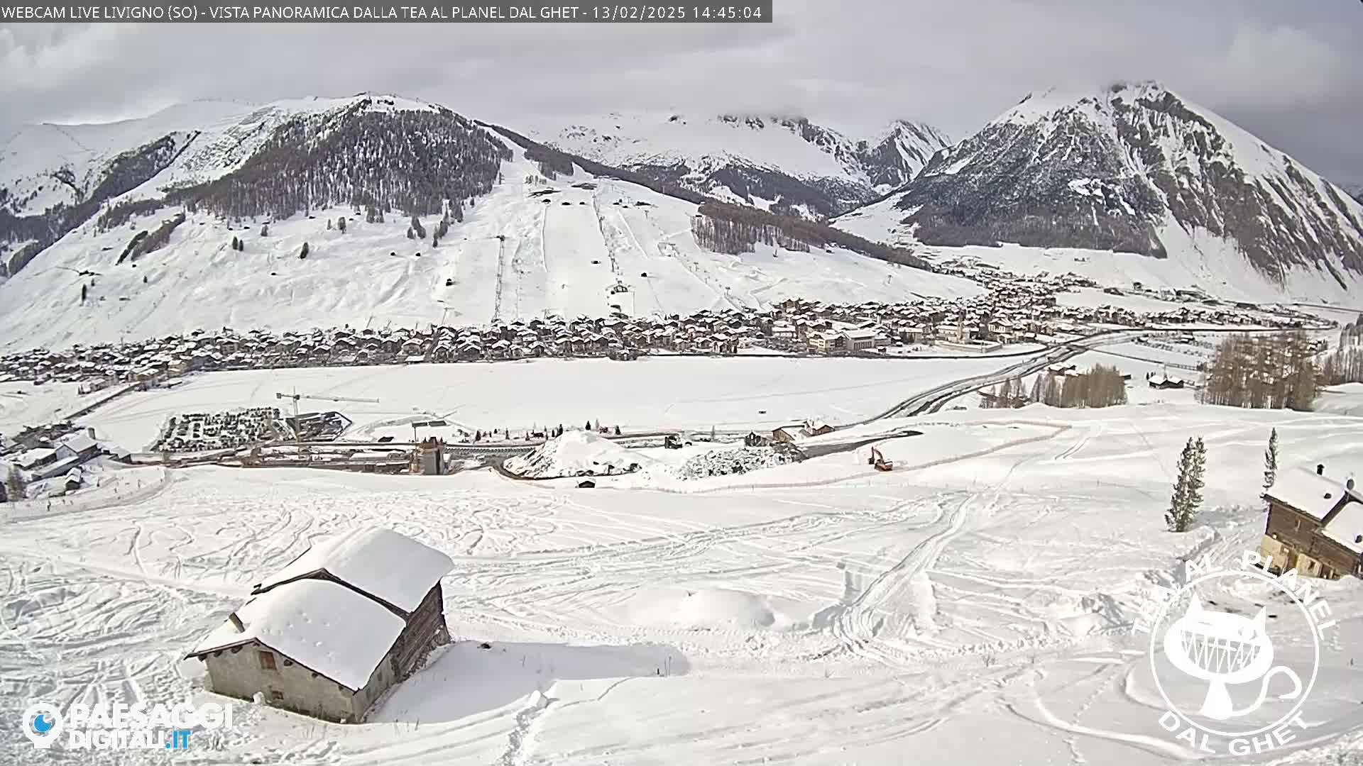 A snow-covered mountain village is nestled in a valley, with snow-covered slopes and mountains in the background under a bright, mostly clear sky.