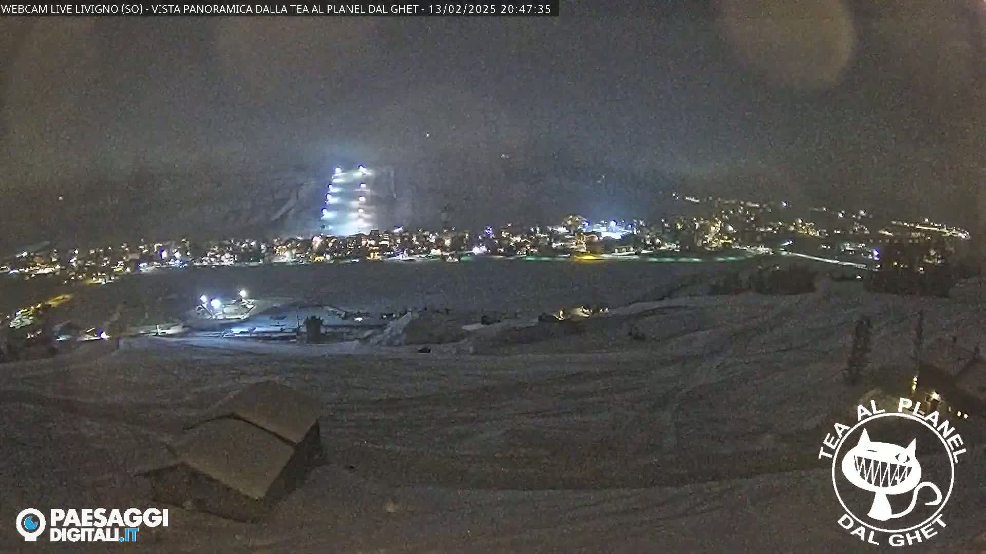 A nighttime, snowy scene shows a village illuminated below, with a brightly lit structure on a hillside, under a dark, possibly overcast sky.