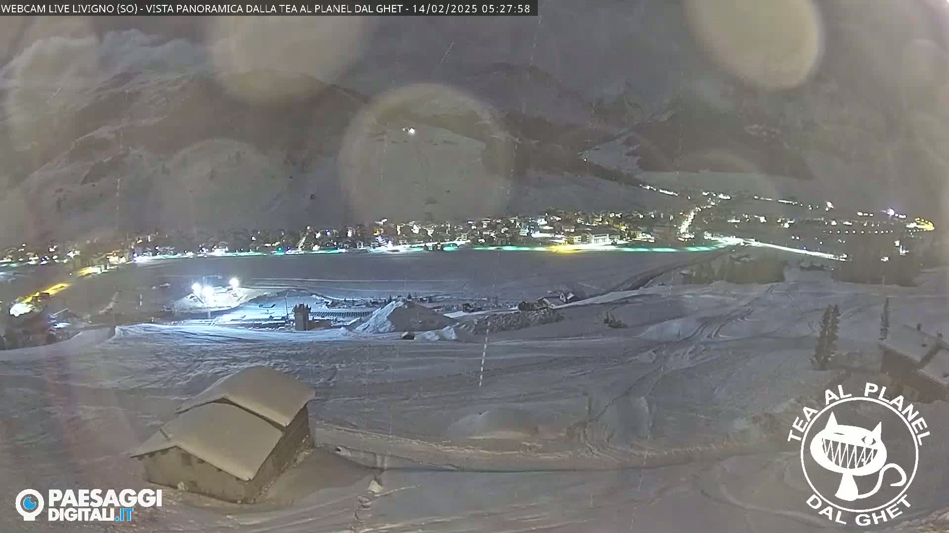 A nighttime, snow-covered landscape shows a village illuminated below, viewed from a higher vantage point with a small snow-covered building in the foreground.