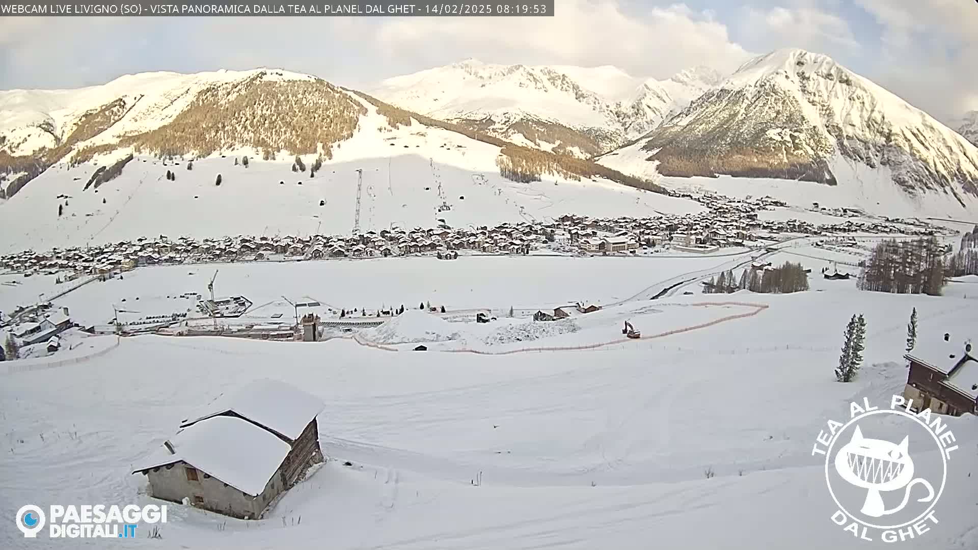 A snow-covered village nestled in a valley between snow-capped mountains under a partly cloudy sky.