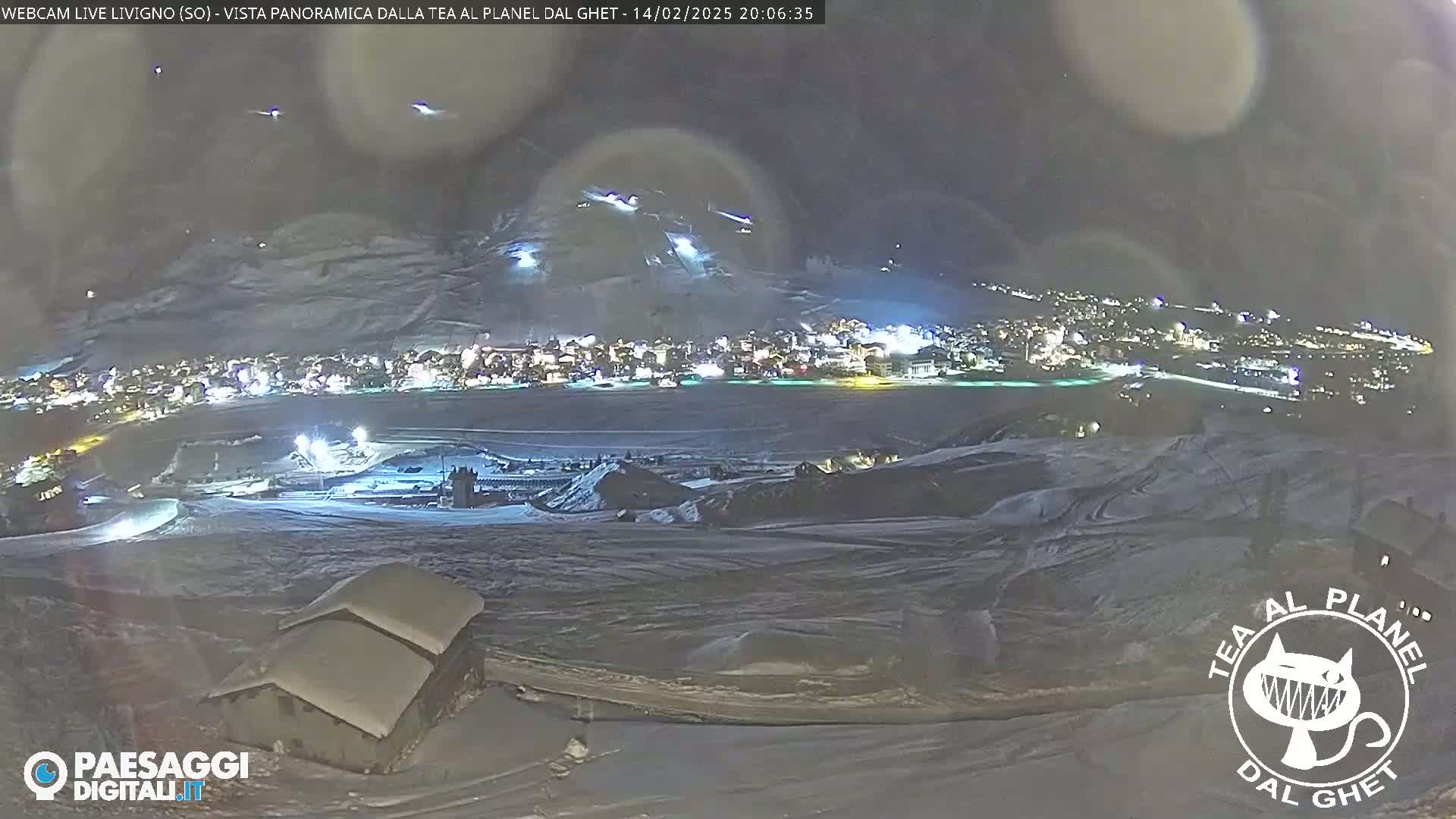 A nighttime, snowy landscape shows a brightly lit town nestled in a valley below a snow-covered mountain with ski slopes, viewed from a higher vantage point.