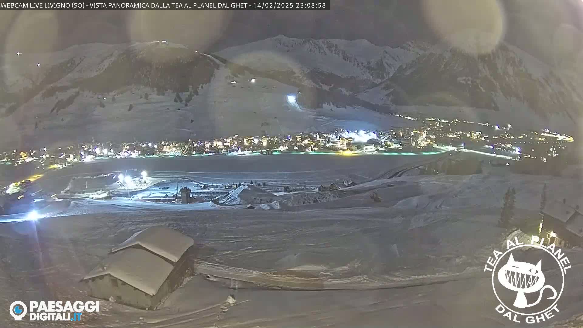 A nighttime, snowy mountain landscape shows a town illuminated below, with a small snow-covered building in the foreground.