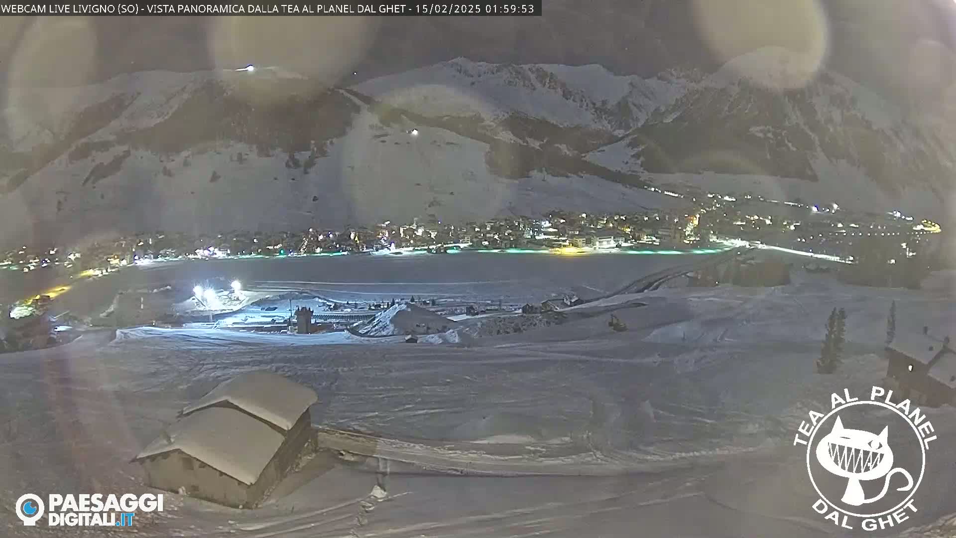 A snow-covered landscape at night, showing a town illuminated in the distance below a snow-capped mountain.