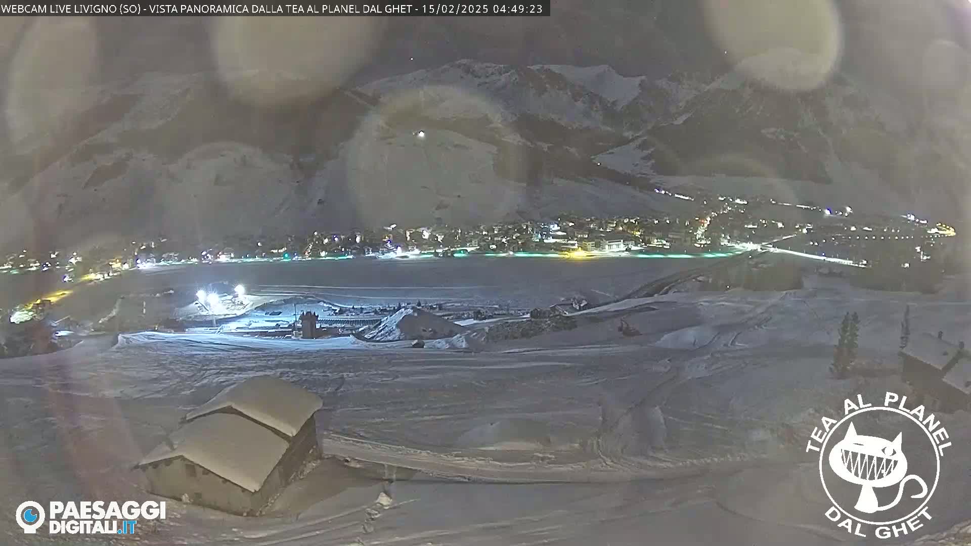 A nighttime, snow-covered landscape shows a village illuminated below, with a small building in the foreground and mountains in the background under a dark, cloudy sky.