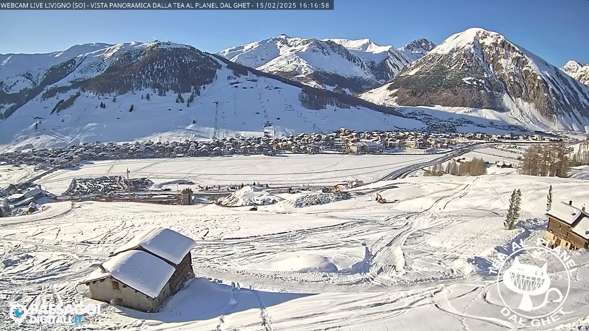 A snow-covered mountain village is seen from a high vantage point on a sunny day, with several small buildings visible in the foreground.