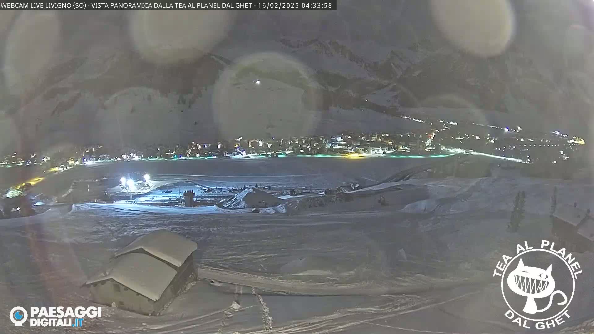 A nighttime, snowy, mountainous landscape shows a village in the distance, illuminated, with a snow-covered building in the foreground under a hazy sky.