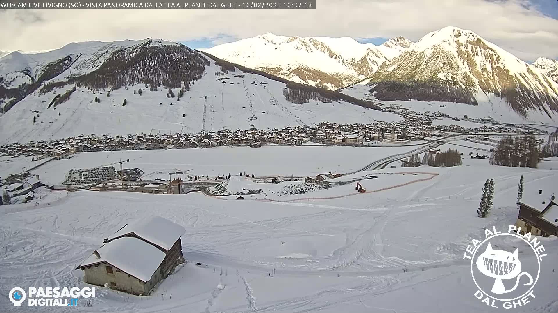 A snow-covered mountain village is visible from a snowy hillside, with partly cloudy skies overhead.