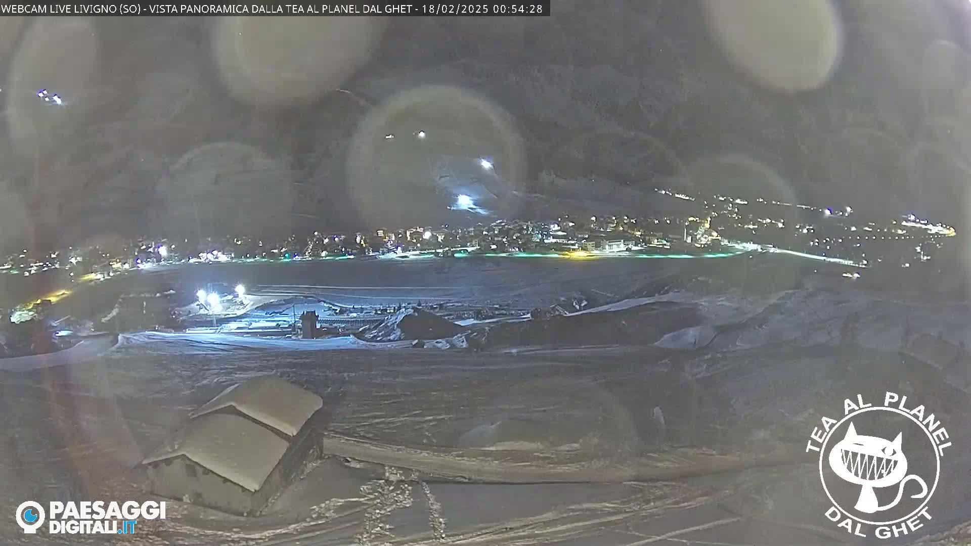 A nighttime, snowy landscape shows a town in the distance illuminated by lights, with a few buildings in the foreground under a hazy, dark sky.