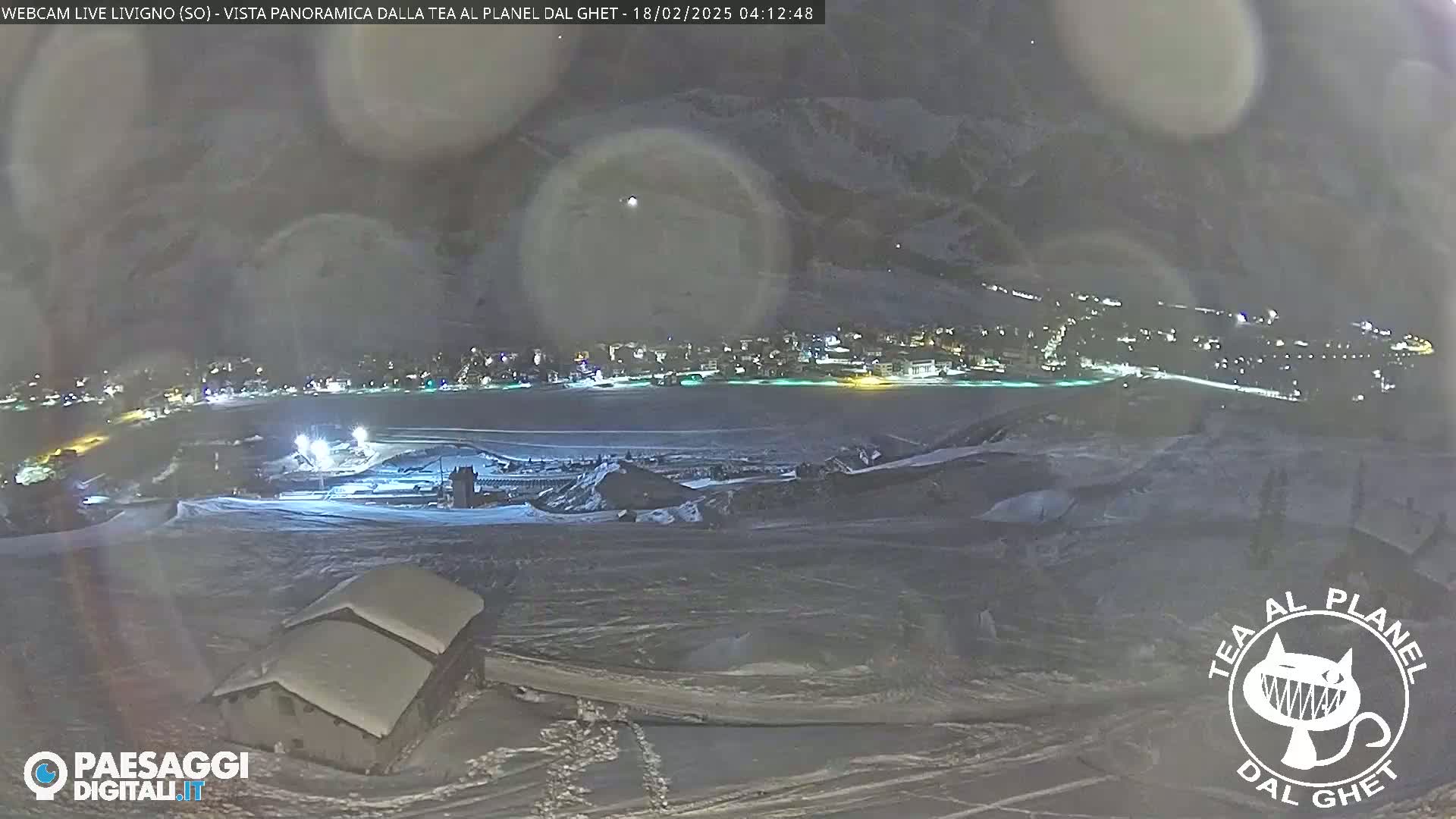 A nighttime, snowy panorama shows a town illuminated below, with a few buildings in the foreground and mountains in the background under a hazy sky.