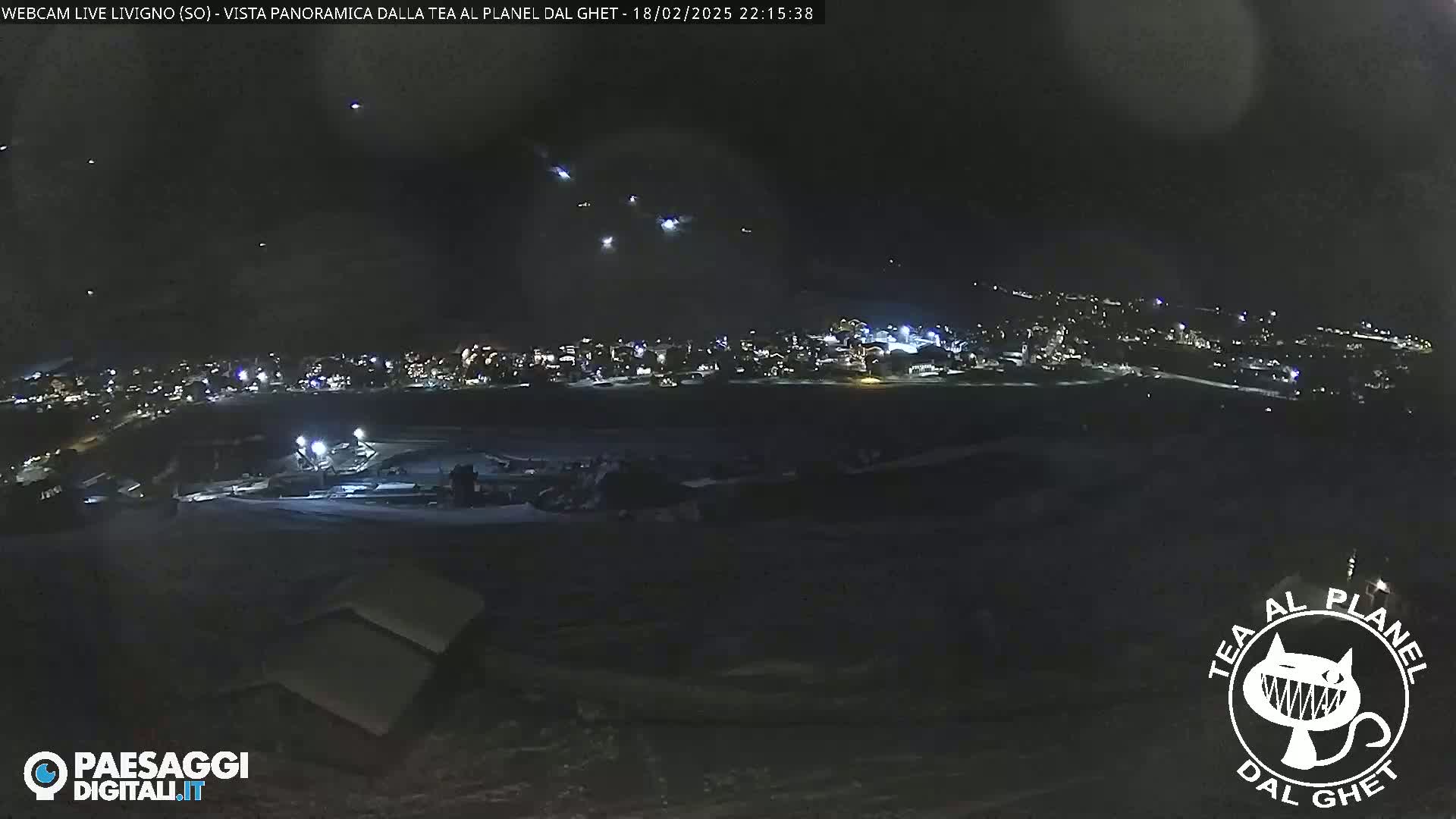 A nighttime, panoramic view of a snow-covered town illuminated by lights, with several faint lights in the sky above.