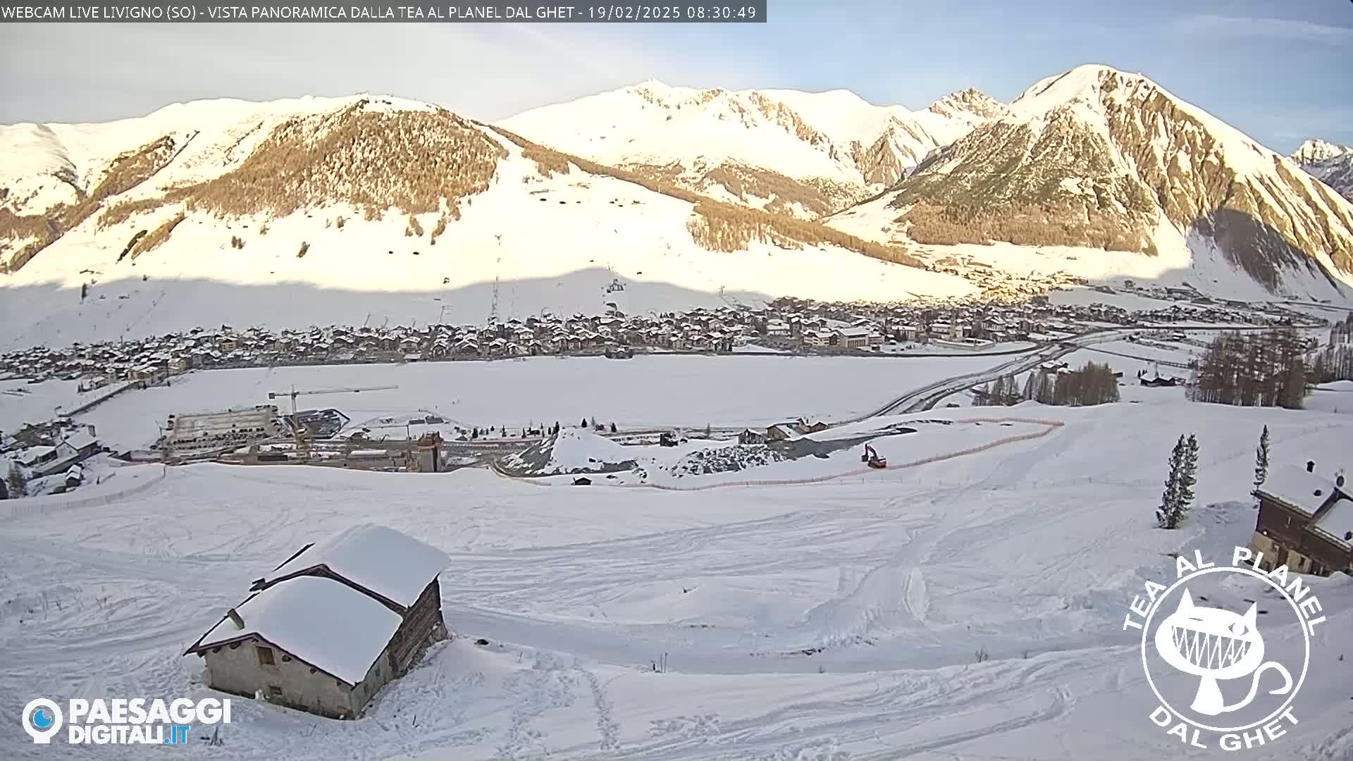 A snow-covered landscape shows a village nestled in a valley between snow-capped mountains under a clear, sunny sky.