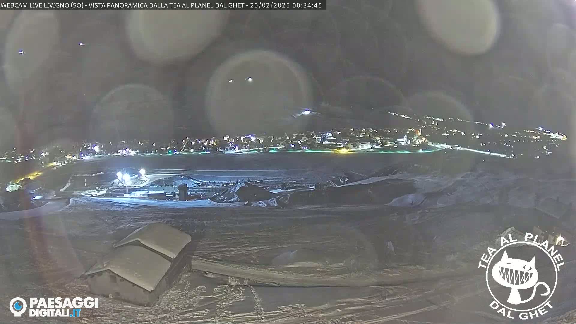 A nighttime, snowy landscape shows a town illuminated in the distance, with two snow-covered buildings in the foreground under a dark, possibly overcast sky.