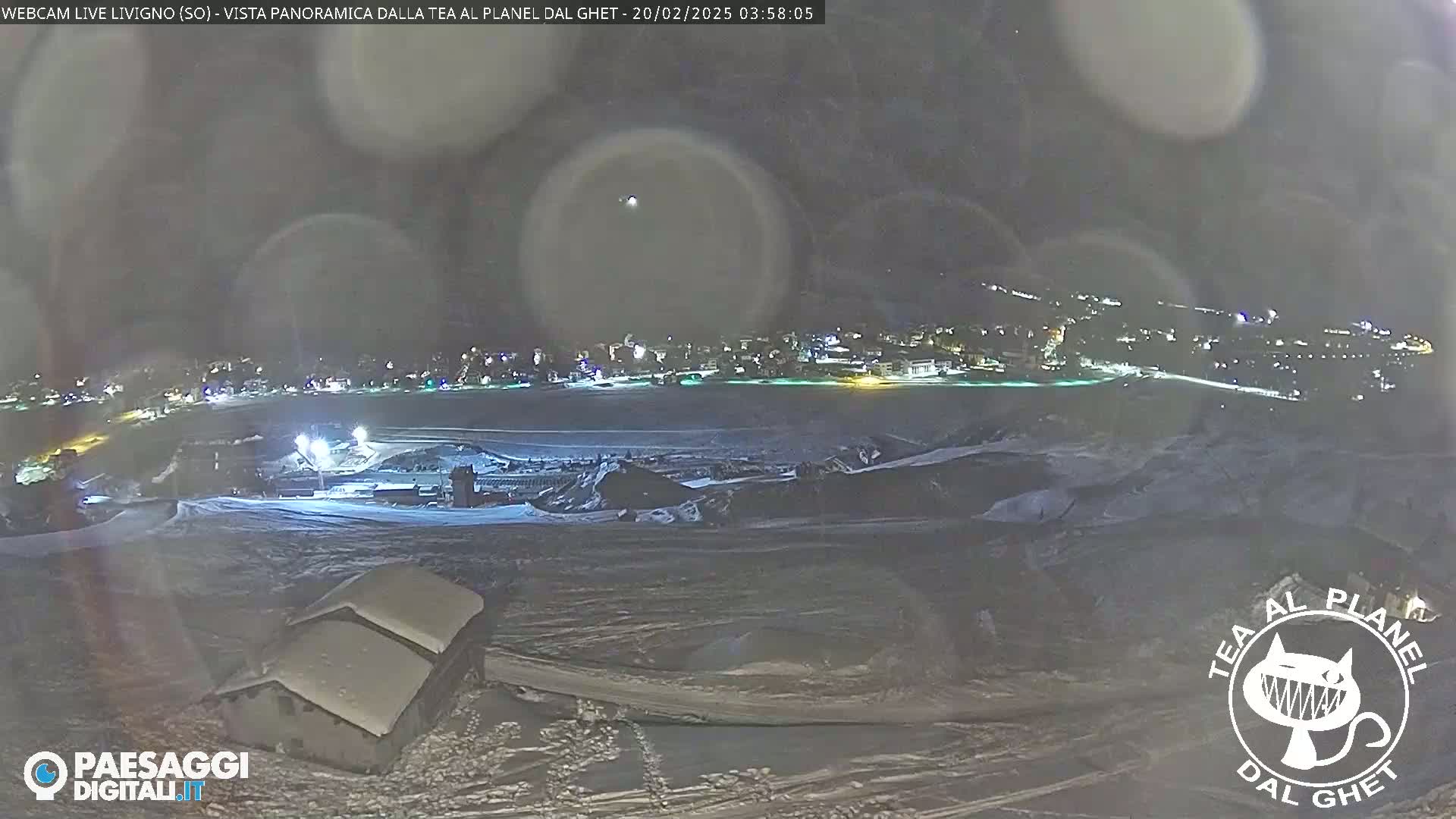 A nighttime, snowy panorama shows a village lit up in the distance, with a closer view of snow-covered buildings and land.