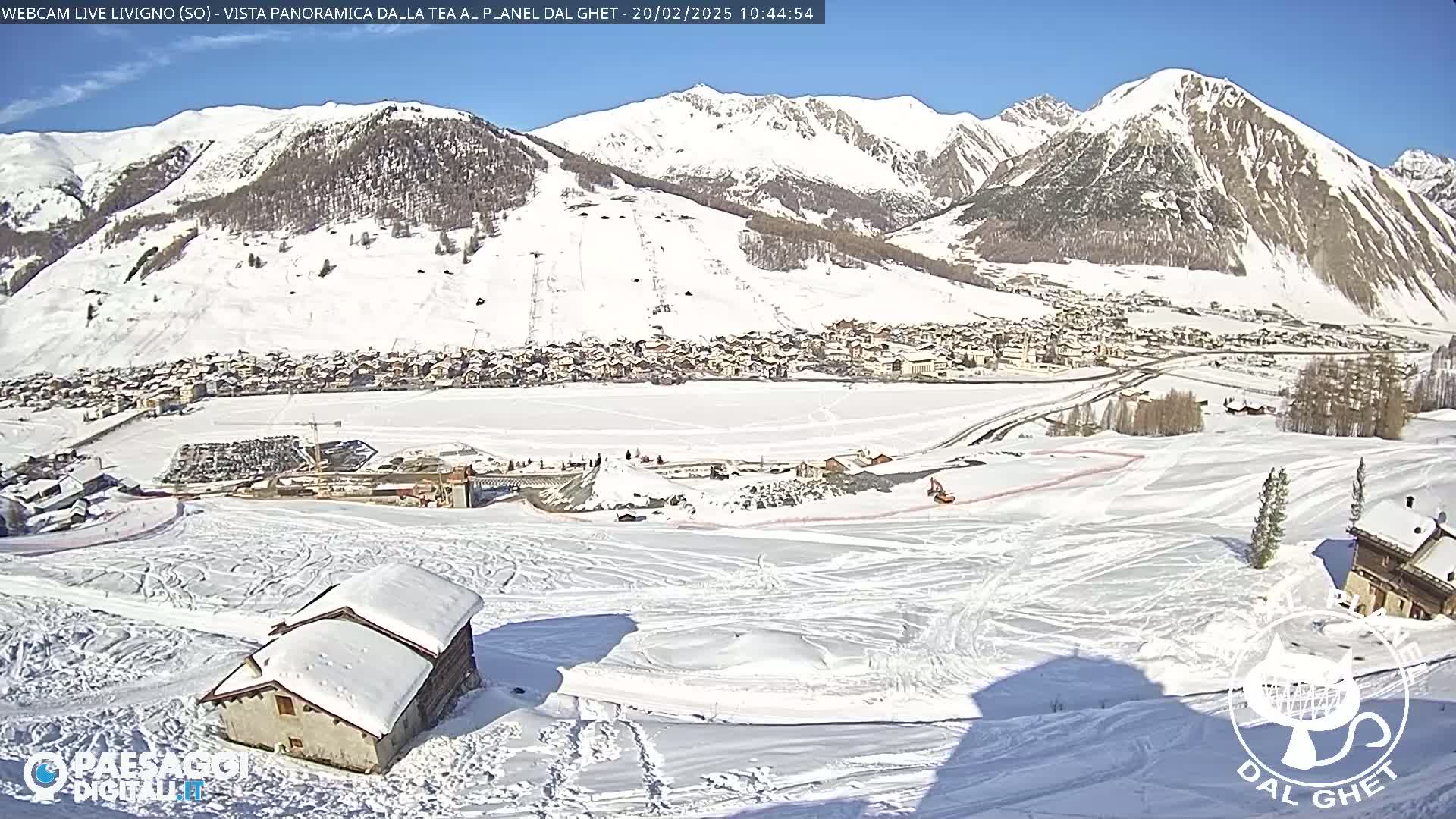 A snow-covered mountain village is seen from a high vantage point on a sunny day, with snow-covered slopes and a small building in the foreground.