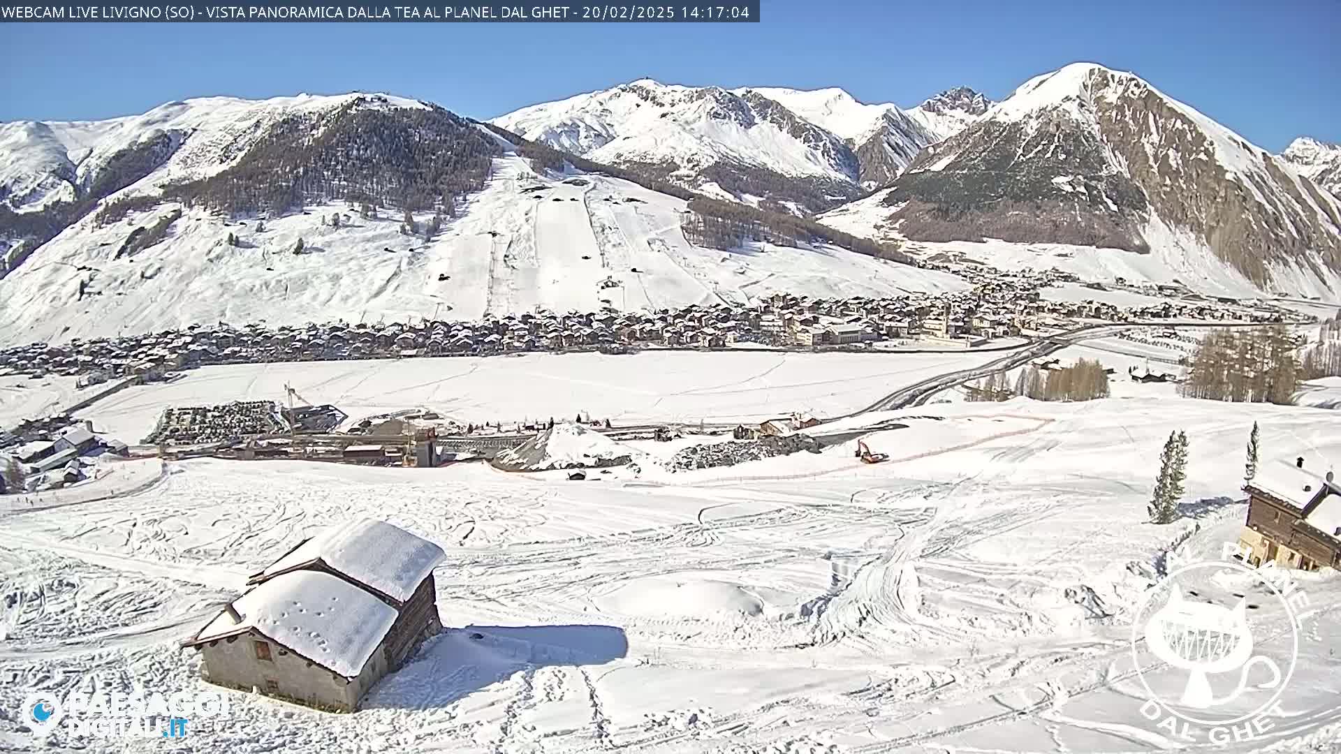 A snow-covered mountain village is seen from a high vantage point on a sunny day, with a small snow-covered house in the foreground and ski runs visible on the mountainside.