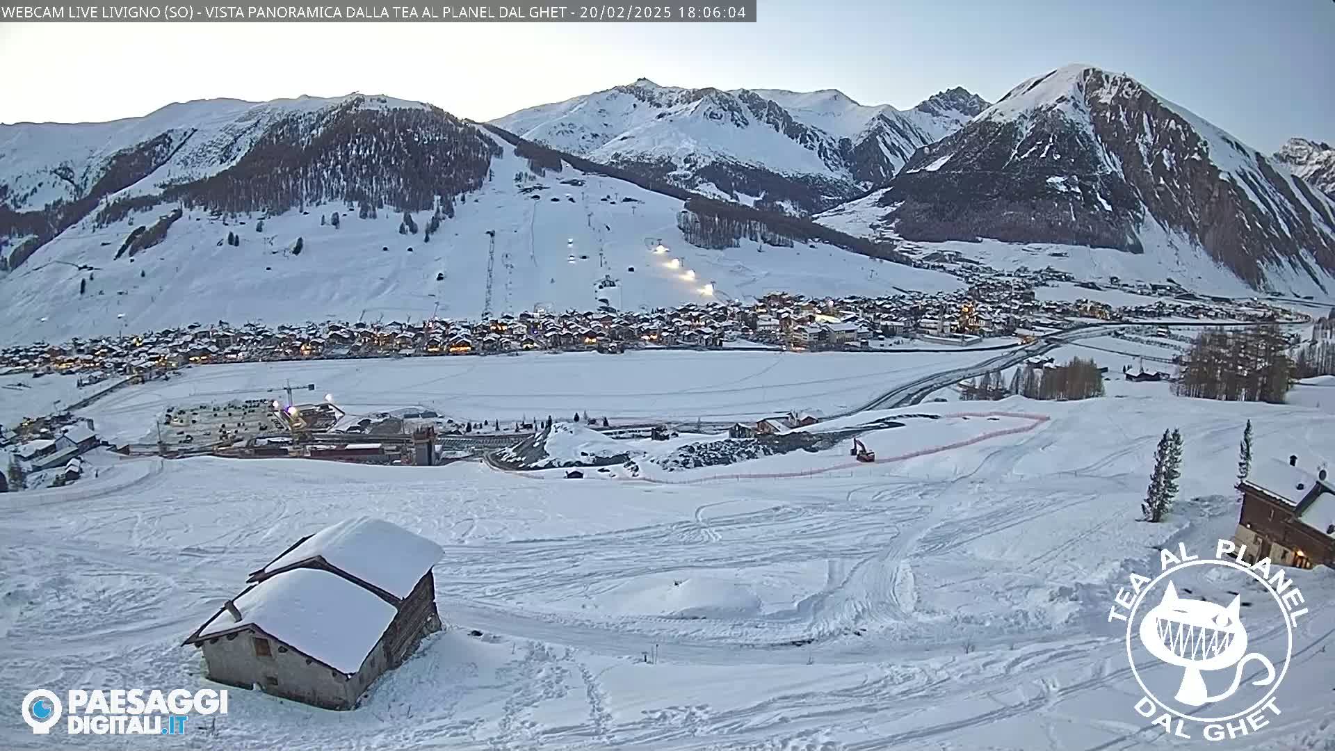A snow-covered mountain village is visible beneath a mostly clear twilight sky, with snow-covered mountains in the background and a small snow-covered building in the foreground.