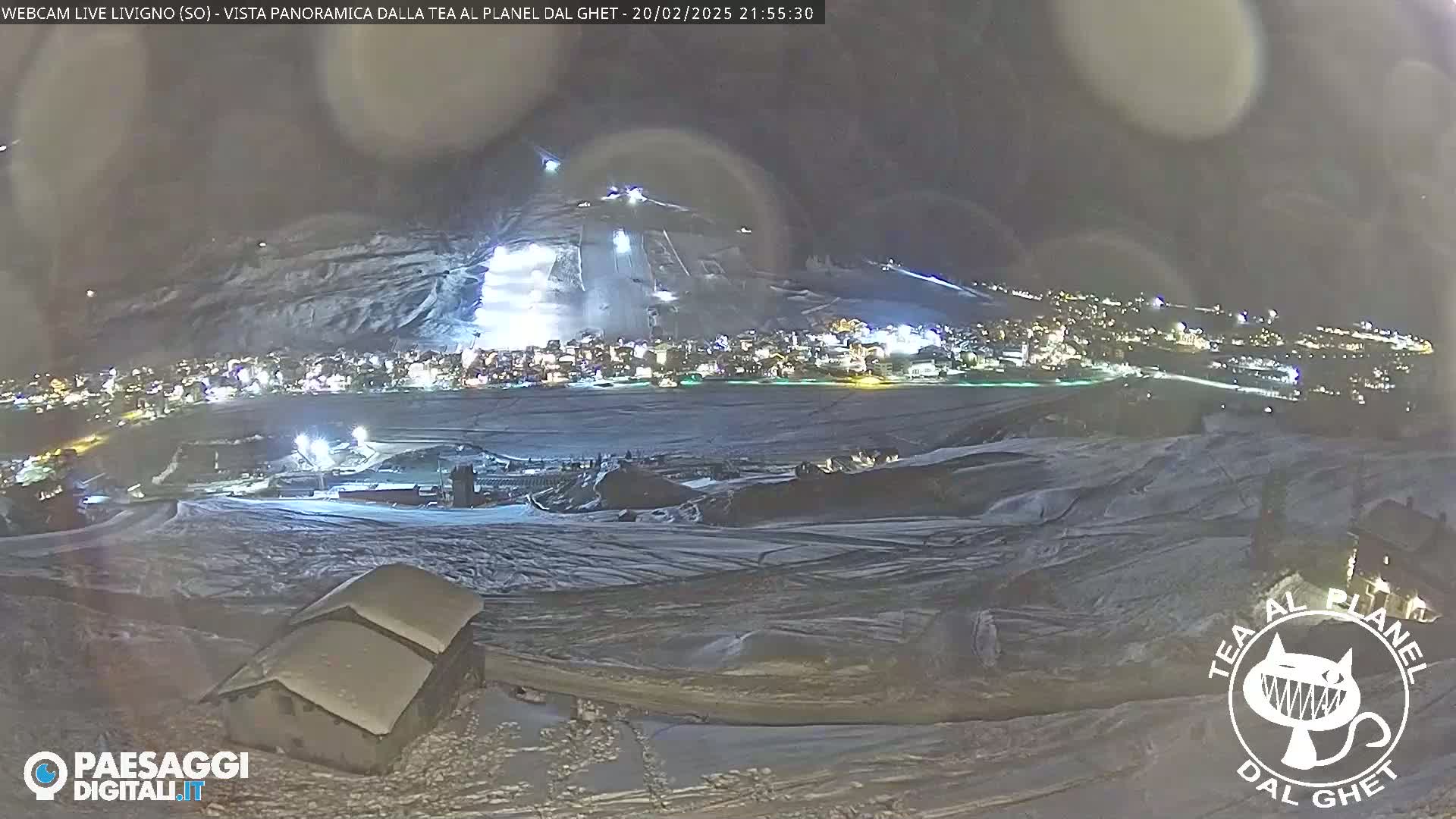 A nighttime, snow-covered landscape shows a town illuminated below a hill with lighted ski slopes, under a dark, slightly hazy sky.