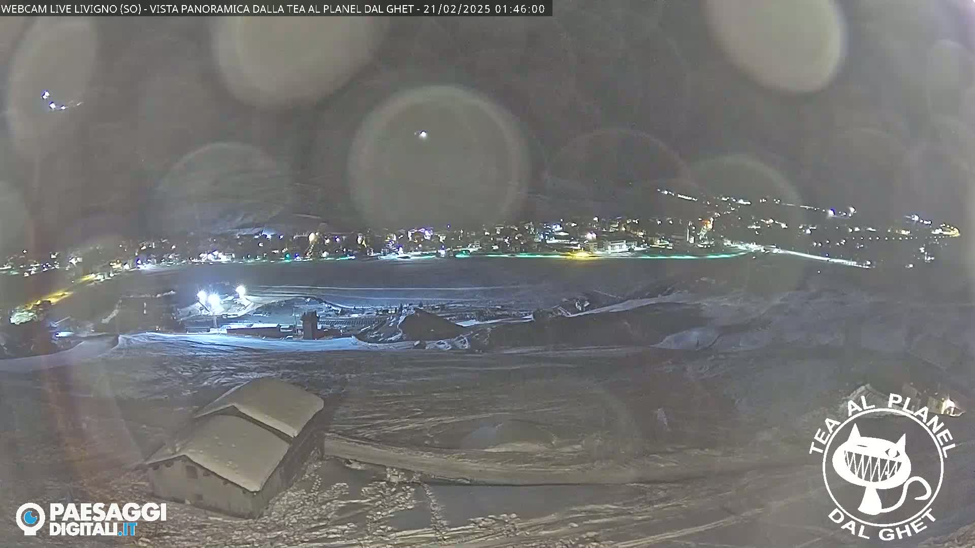 A nighttime, snowy, panoramic view of a town illuminated by lights, with a few buildings in the foreground under a hazy sky.