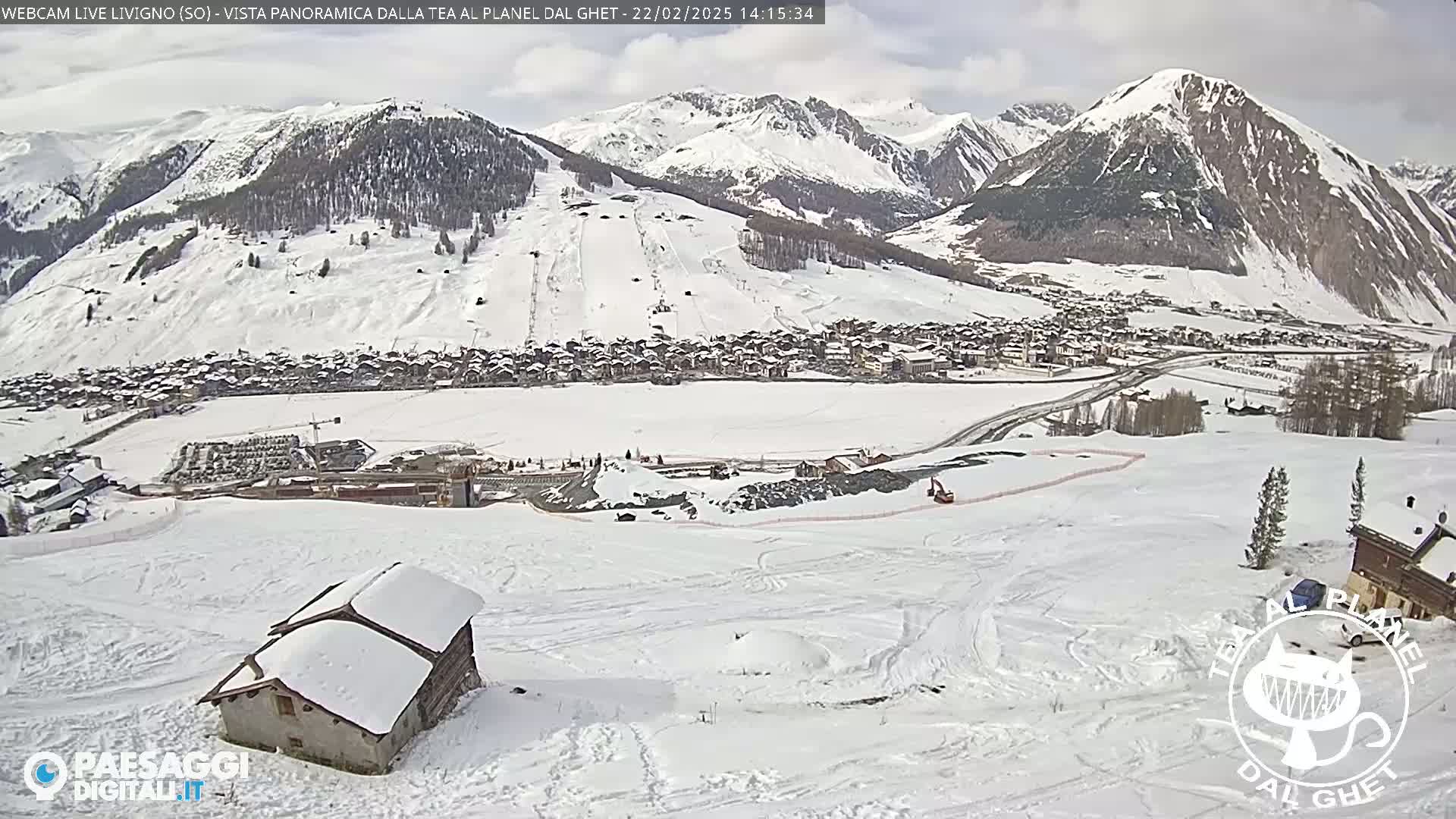 A snow-covered mountain village is nestled in a valley below snow-covered mountains under a partly cloudy sky.