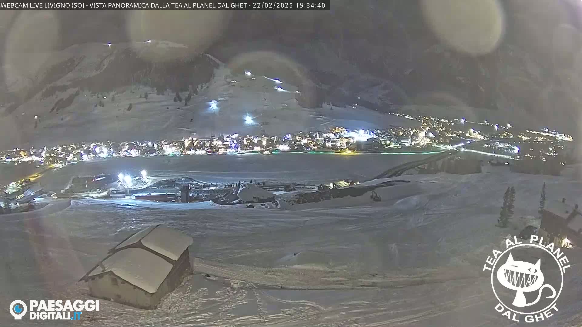 A nighttime view of a snow-covered town nestled at the base of a mountain, illuminated by lights, under a cloudy sky.