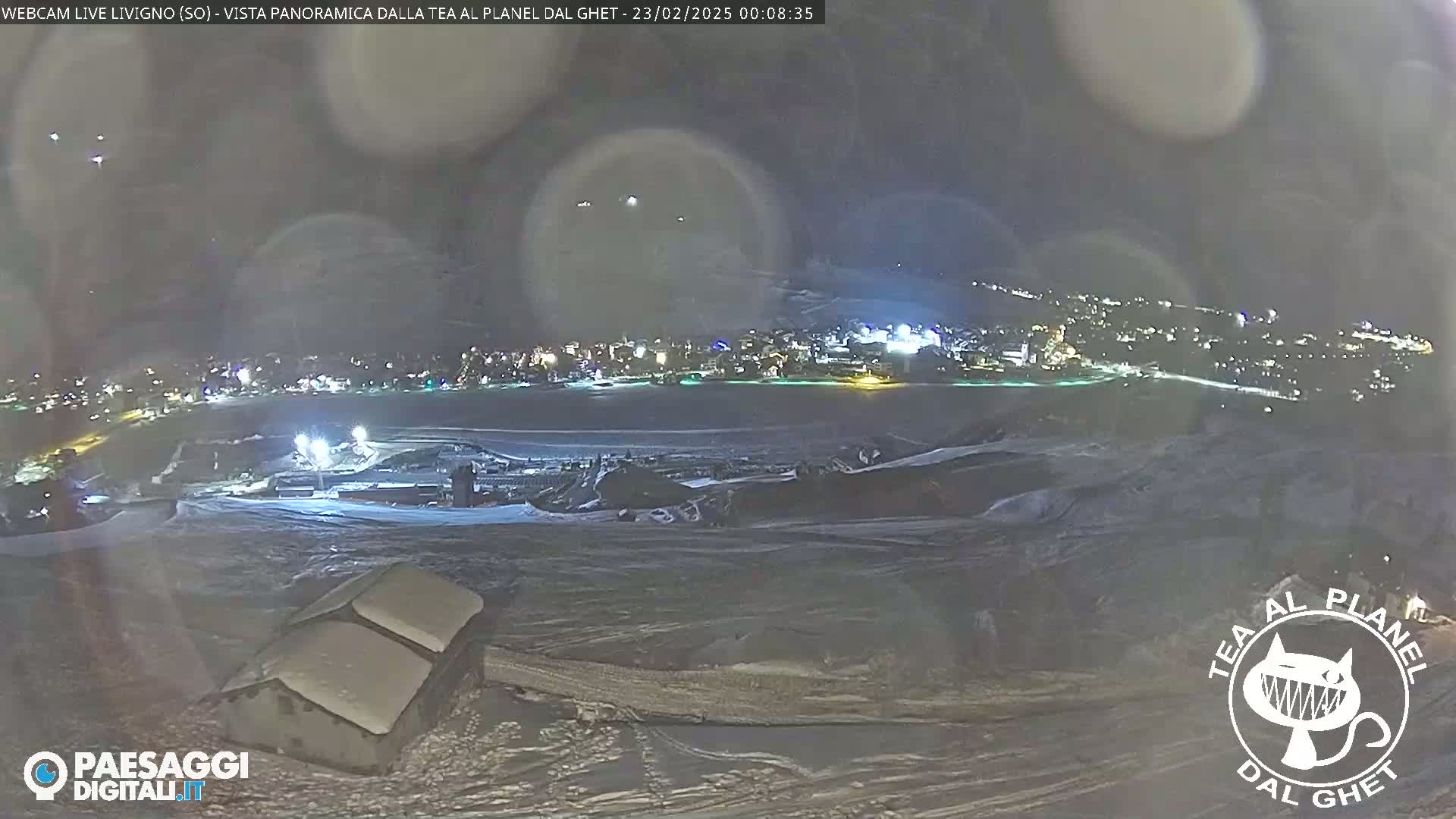 A nighttime, snowy panoramic view of a town illuminated with lights, seen from a higher vantage point overlooking a snow-covered landscape.